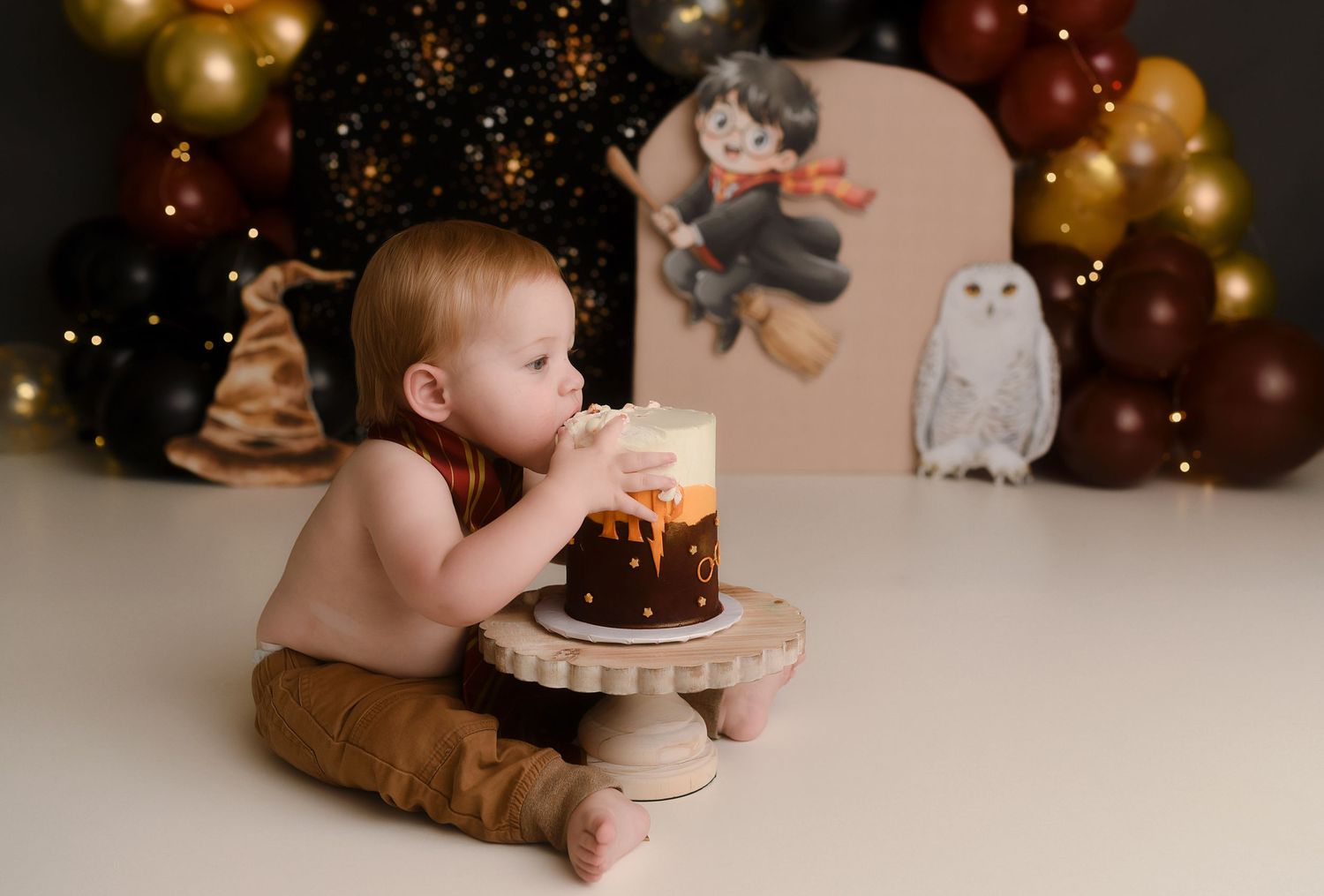 Baby in Harry Potter outfit eating smash cake at themed first birthday photo session with balloon backdrop.