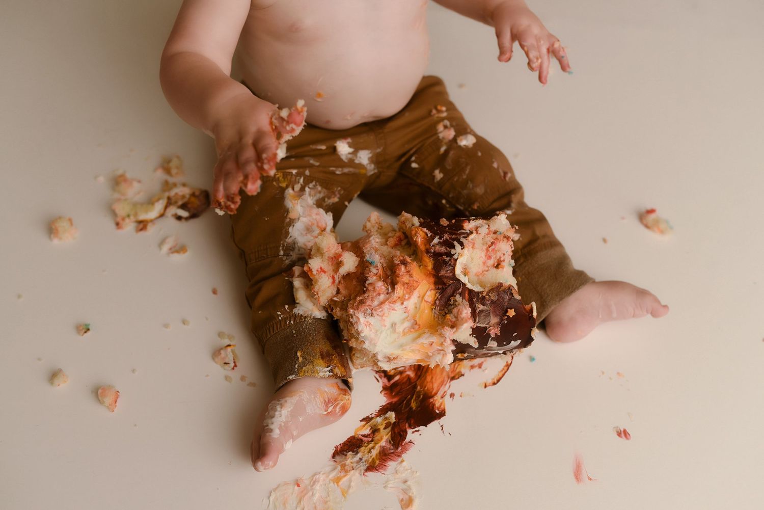 Baby in brown pants sits surrounded by smashed birthday cake during first birthday cake smash photoshoot.