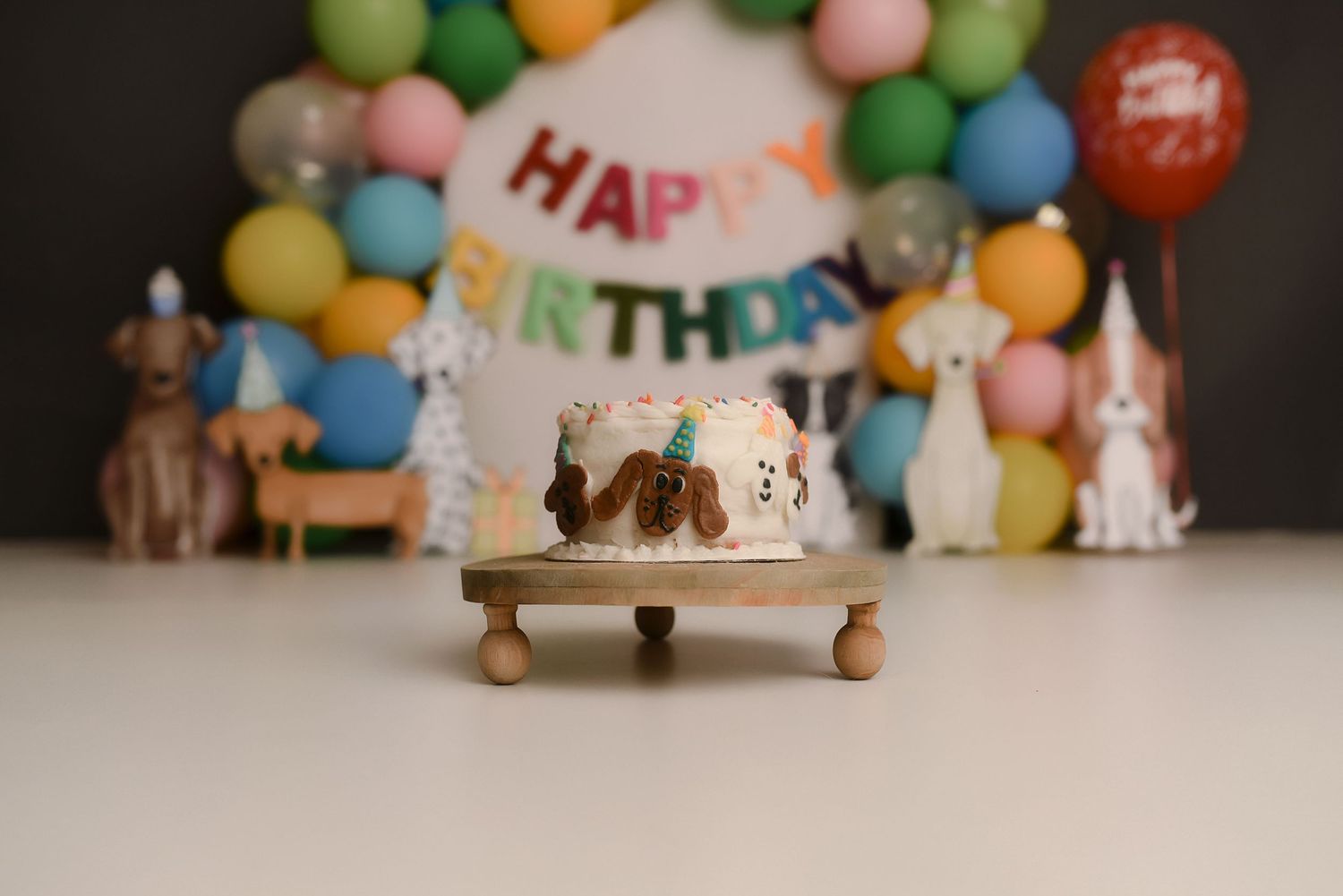 Happy birthday cake on wooden stand with colorful balloon arch and dog figurines in background.