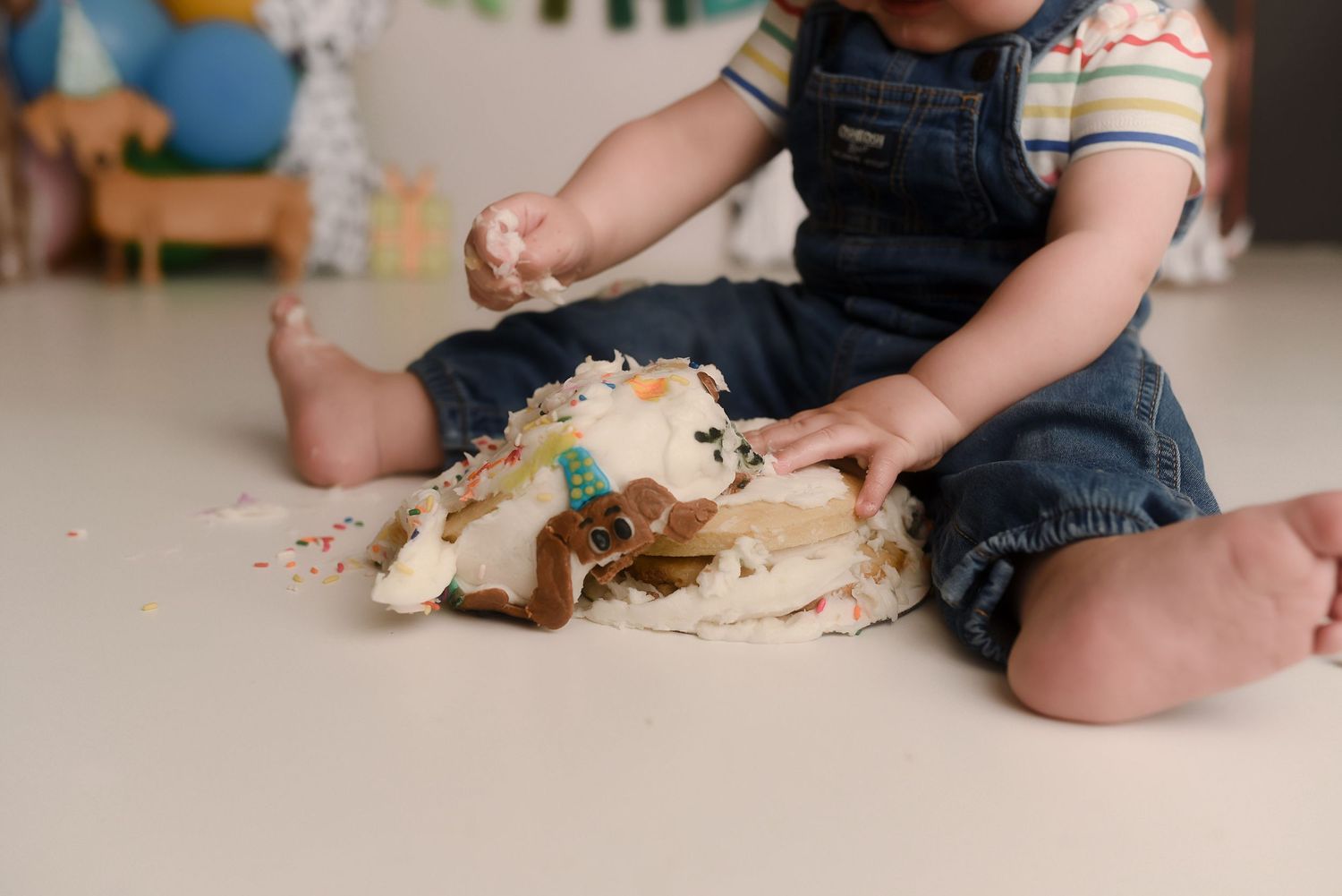 Baby in denim overalls smashing a first birthday cake on a white floor during a cake smash photoshoot.
