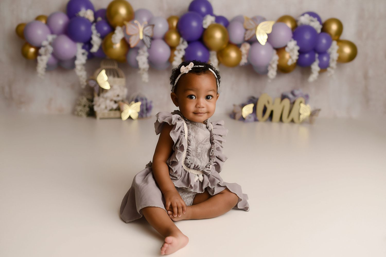 Adorable baby girl in gray ruffle romper sits smiling before purple and gold balloon birthday backdrop.