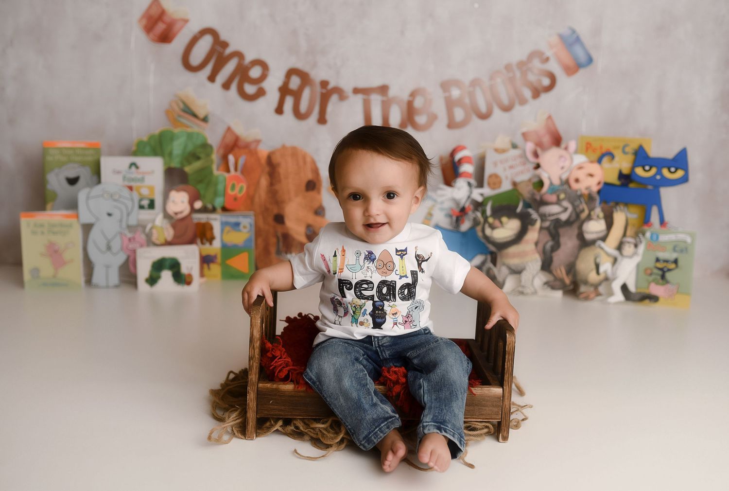 Baby's first birthday photo with books backdrop and One For The Books banner, infant sitting in wooden chair smiling.