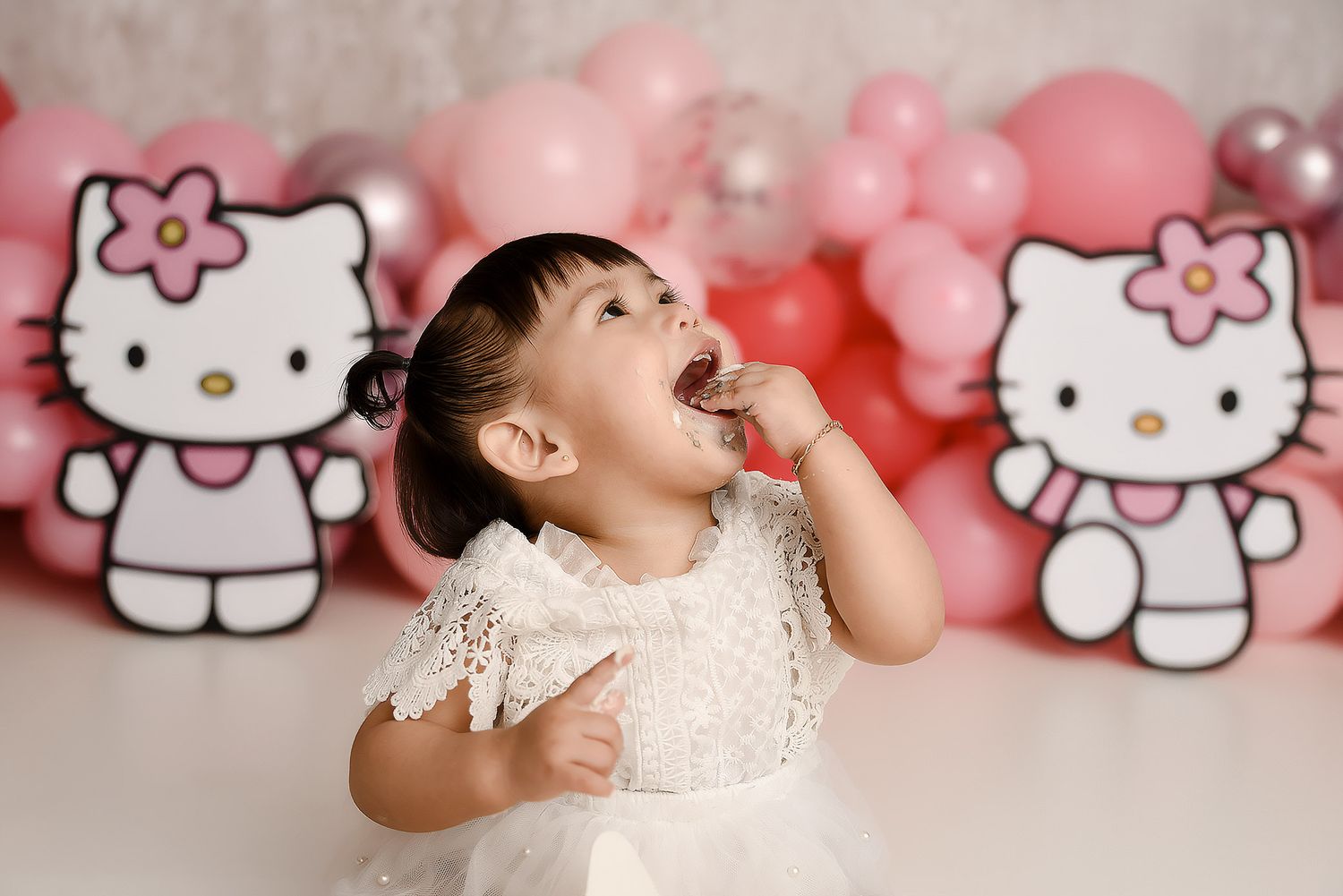 Baby girl in white dress laughing in front of Hello Kitty balloons and pink balloon garland.