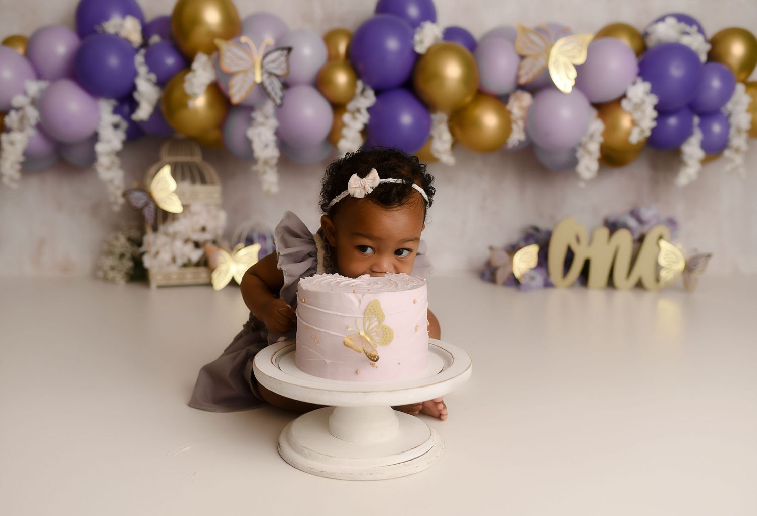 Baby girl in gray dress at purple and gold balloon first birthday cake smash photo session.