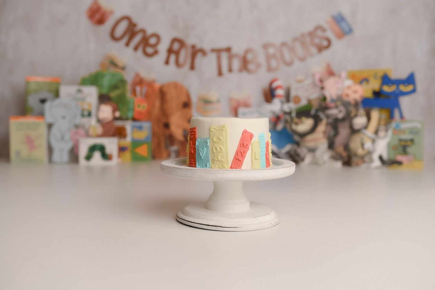 Rainbow-striped first birthday cake on white stand with storybook themed backdrop and colorful children's books.