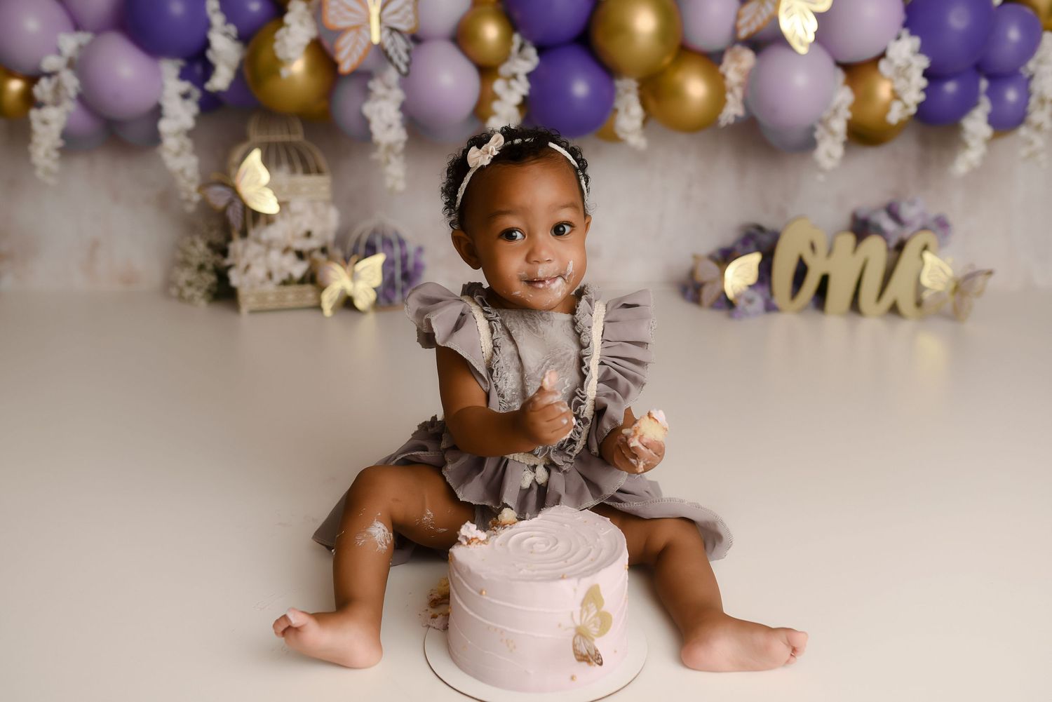 Smiling baby girl in ruffled gray dress sits beside purple butterfly first birthday cake.