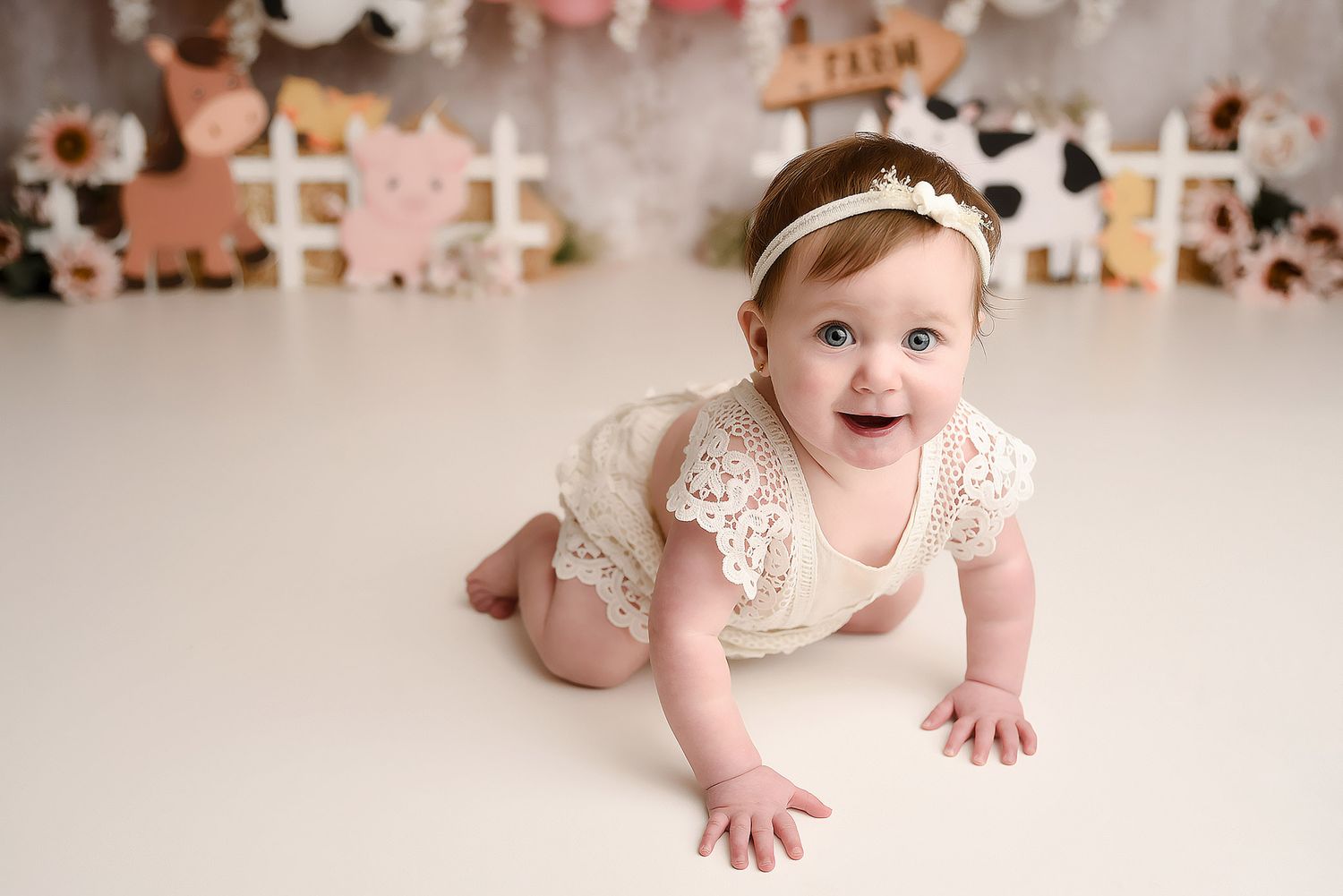 Smiling baby girl in cream lace romper crawling on white floor with farm themed backdrop.