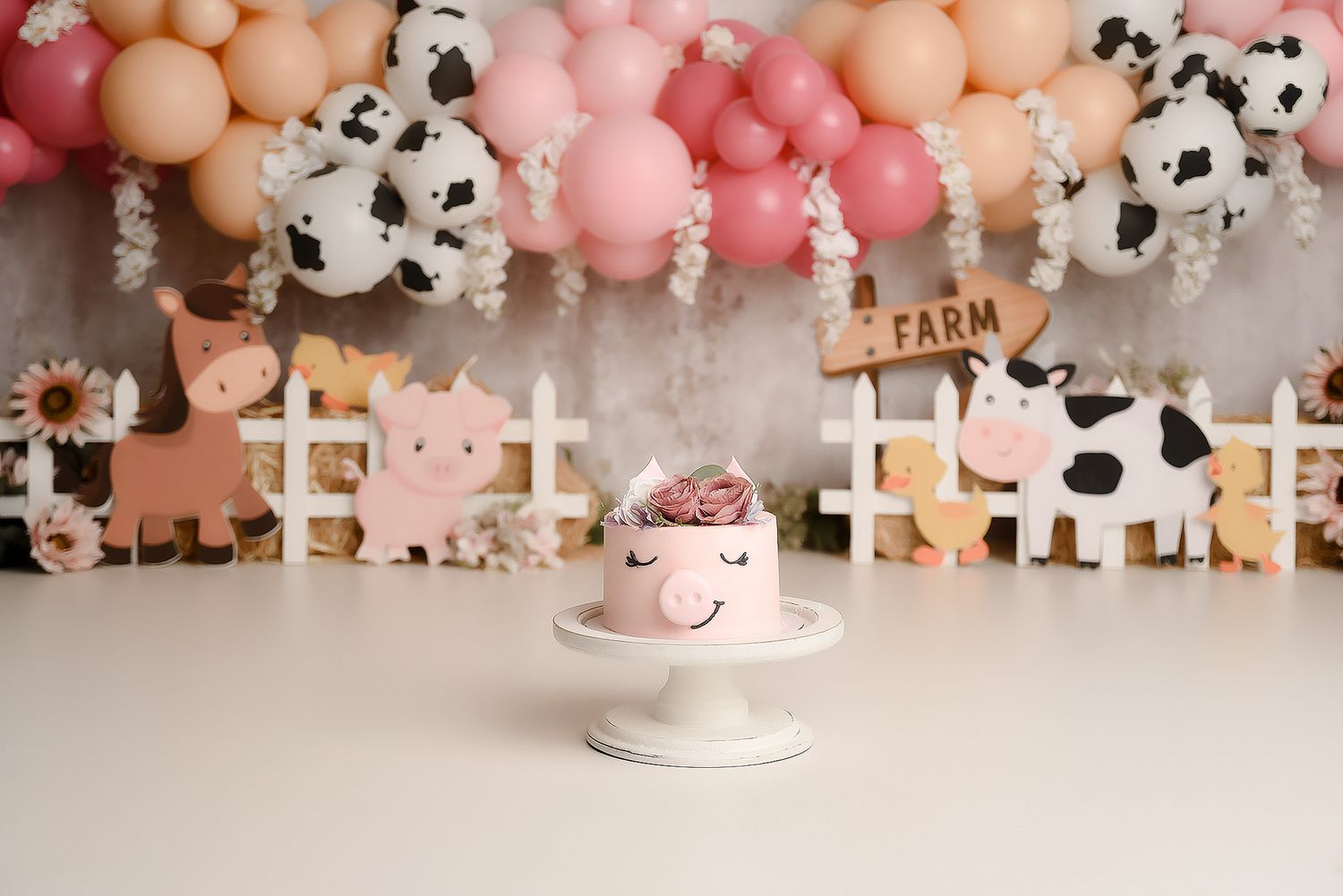 Pink farm-themed smash cake with cow face design on white stand with farm animal decorations.