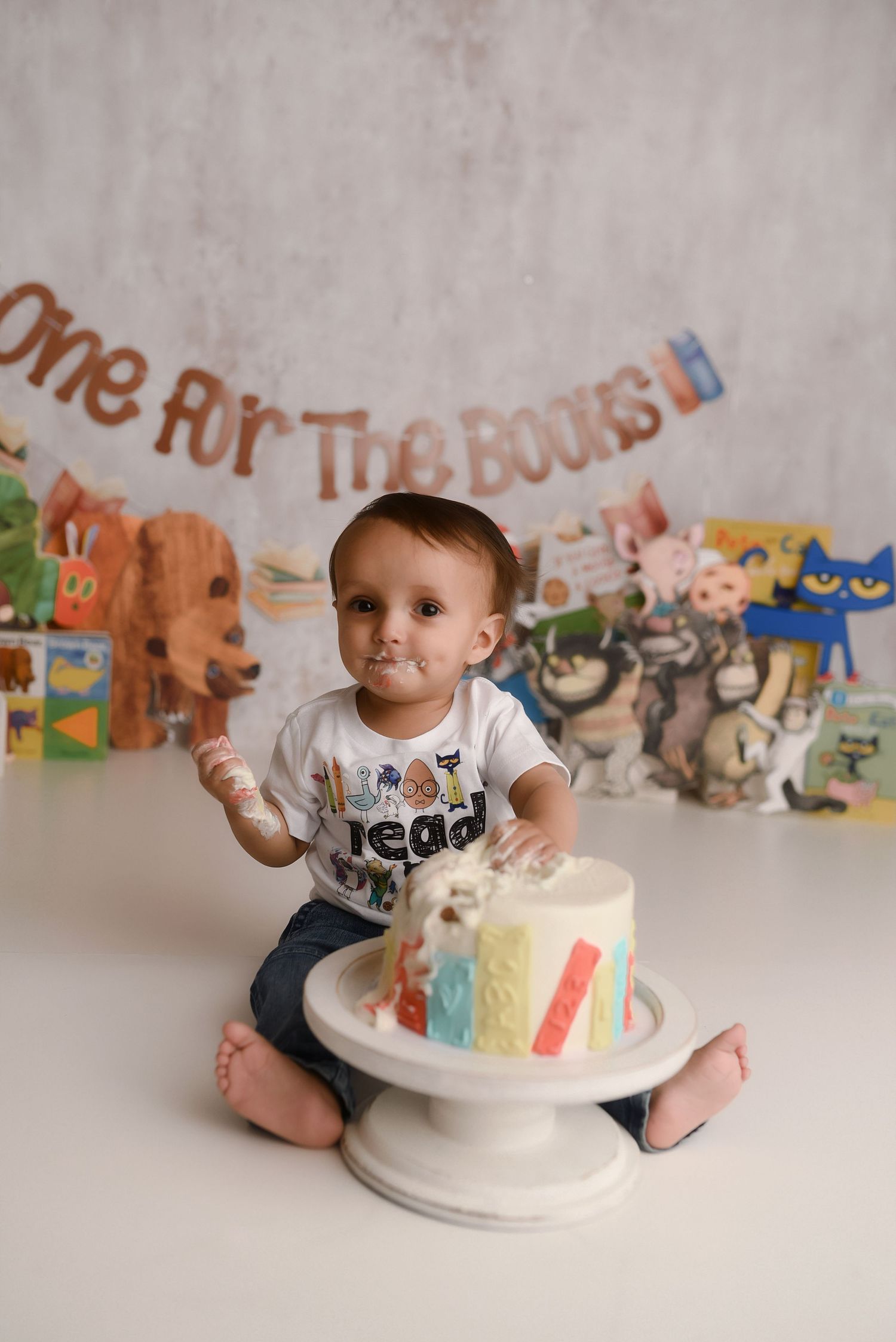 Baby's first birthday cake smash with rainbow cake, children's book themed backdrop, and 'One For The Books' banner.
