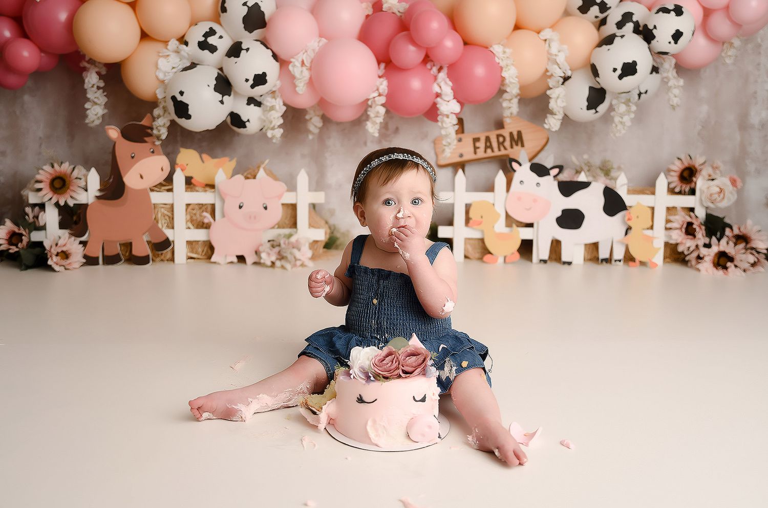 Baby girl in denim outfit smashing pink farm-themed cake surrounded by balloon garland and farm decor.