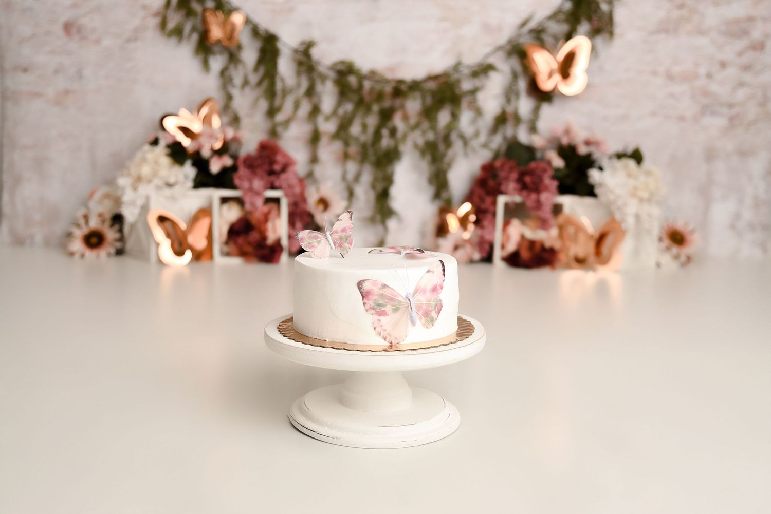 Elegant white butterfly-decorated cake on pedestal with bokeh butterfly lights and floral backdrop.