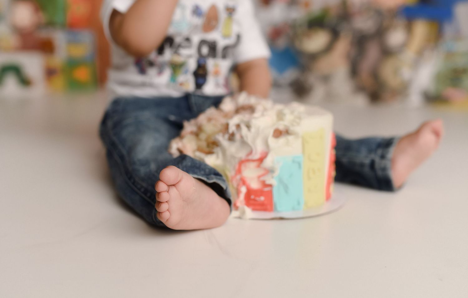 Toddler sitting on floor next to a colorful smash cake during first birthday celebration.