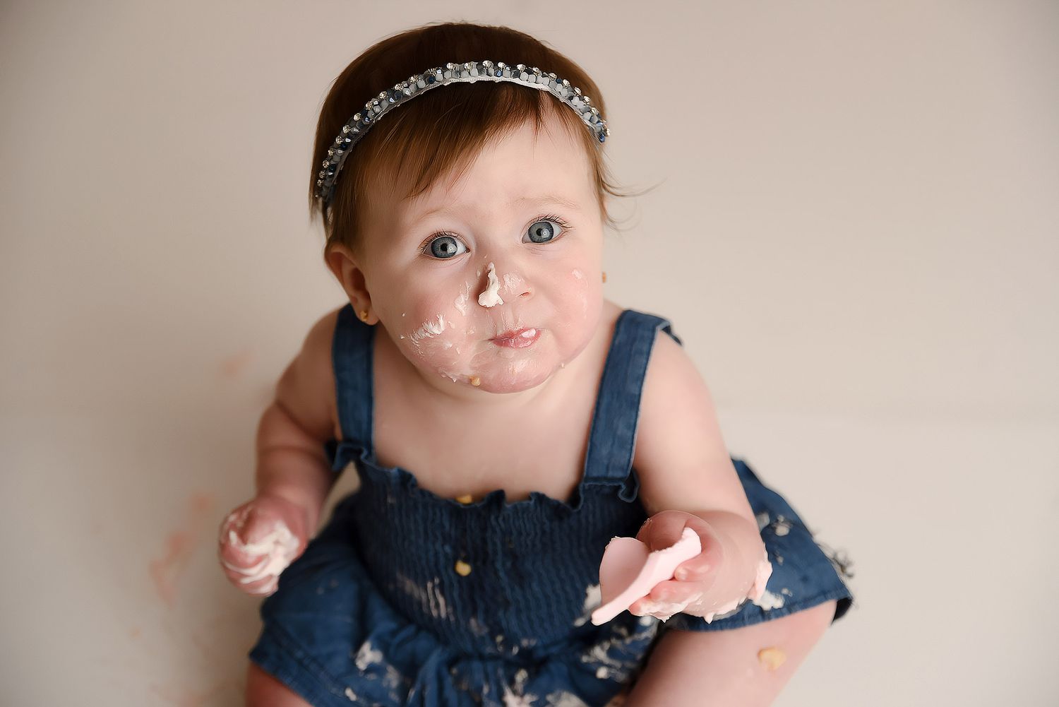 Baby girl in denim dress with cake on her face looking up at camera during smash session.