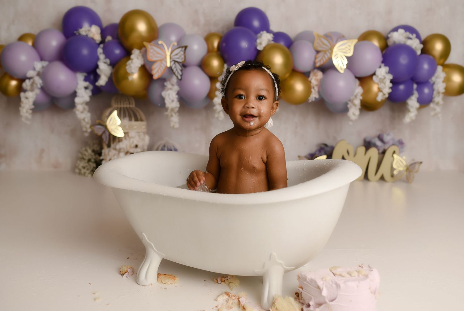 Happy baby girl sits in white clawfoot tub at purple and gold balloon first birthday session.