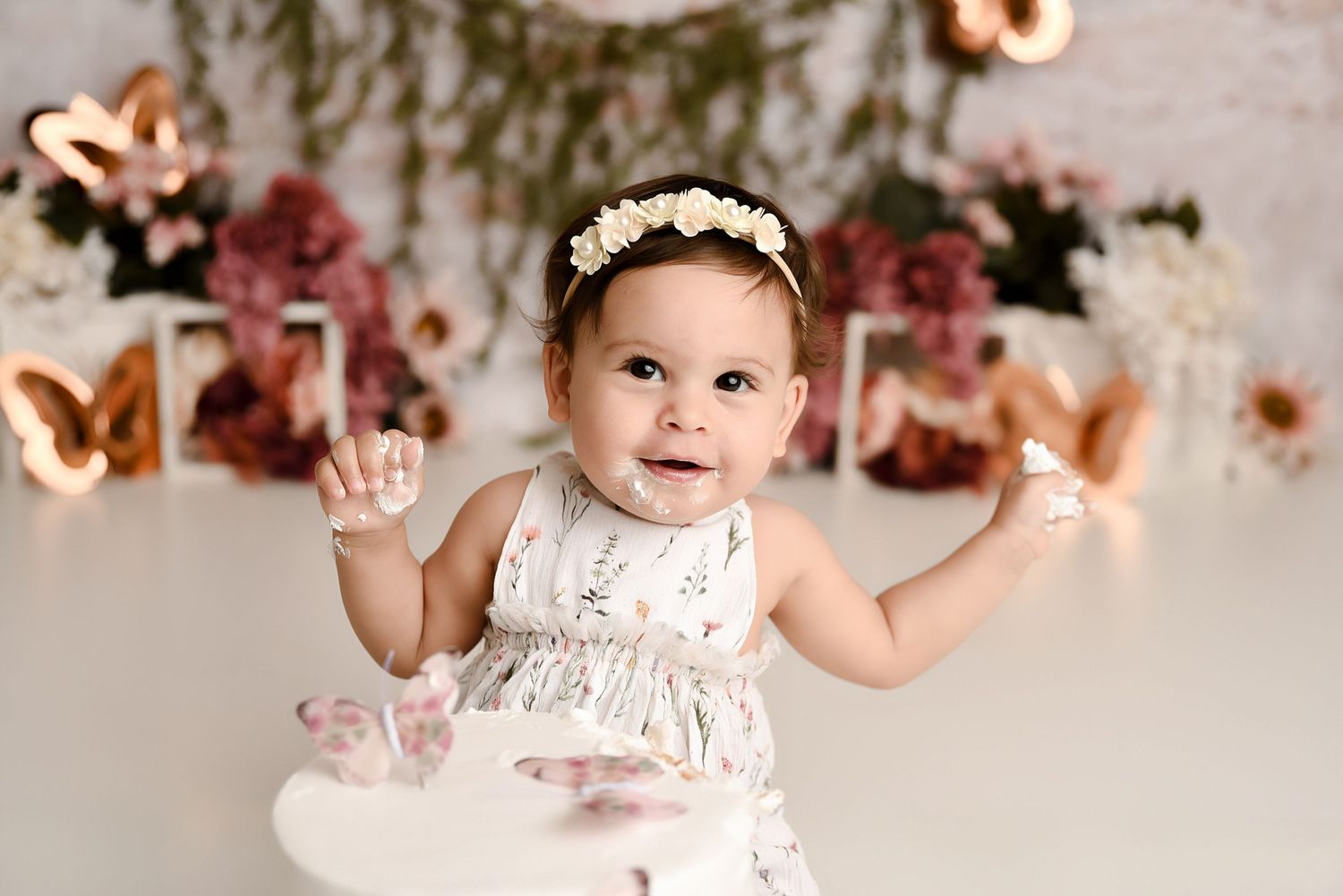 Smiling baby girl in white floral dress with headband celebrating first birthday with cake smash.