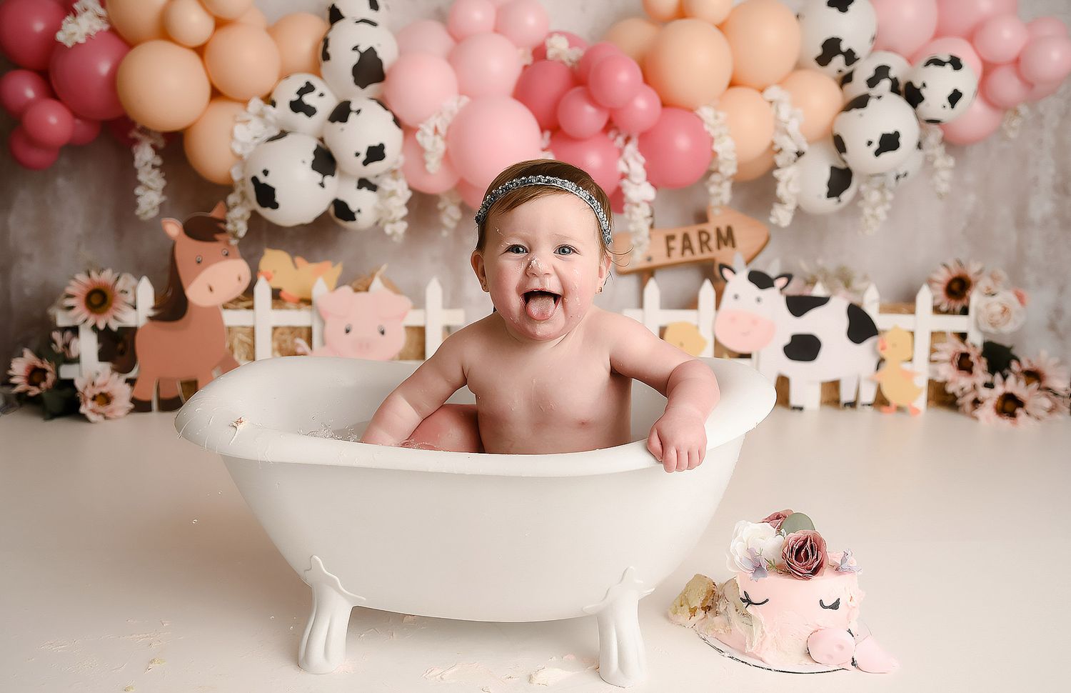 Baby girl sitting in white clawfoot tub laughing with farm themed balloon garland backdrop.
