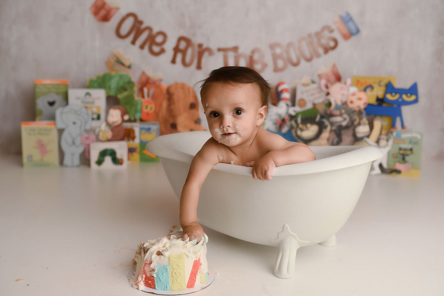 Baby in mini bathtub with smash cake and colorful books, One for the Books banner, first birthday photo session.