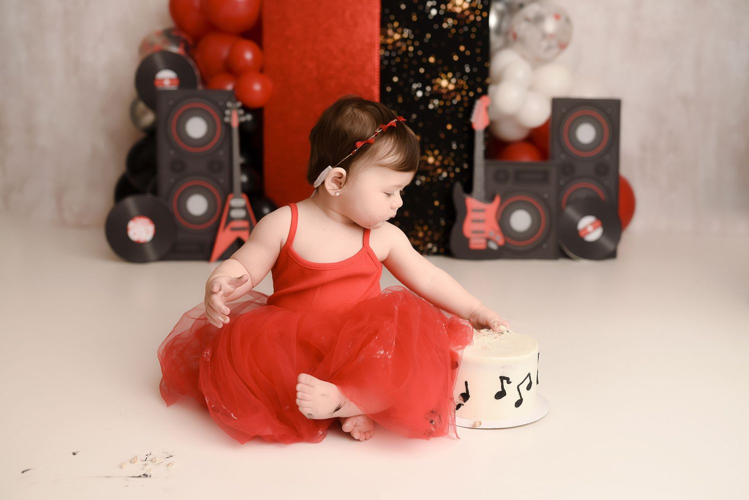 Baby girl in red tutu dress enjoys rock and roll themed cake smash with guitar and speaker backdrop.