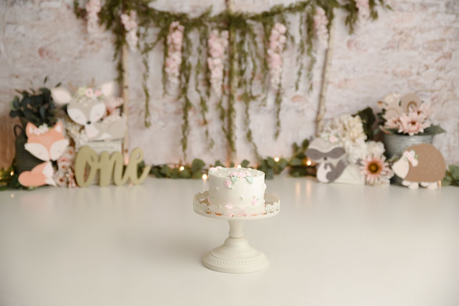 White floral smash cake on mint stand with woodland fox theme and greenery backdrop.