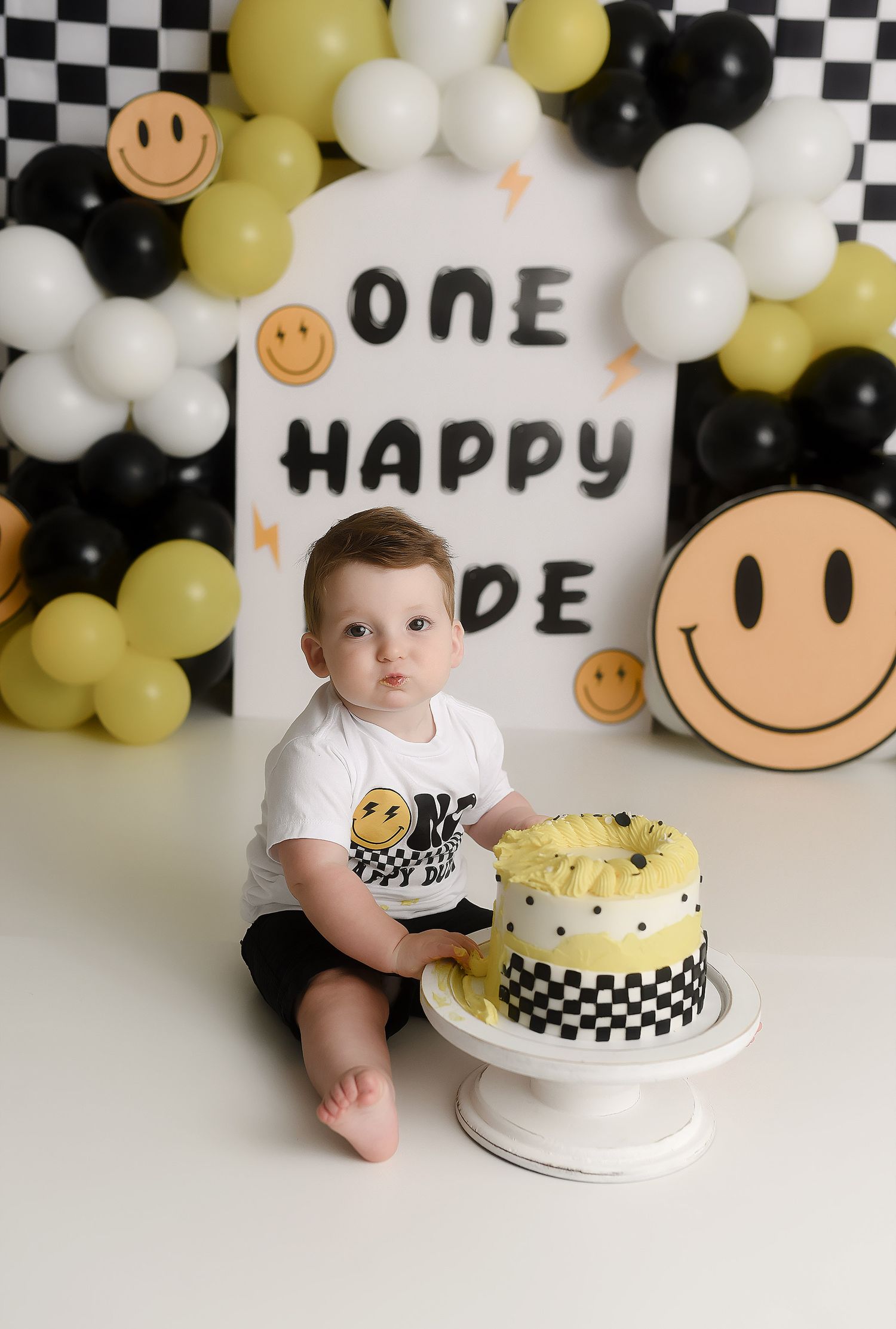 Baby boy first birthday cake smash with smiley face theme, checkered cake, and balloon arch.