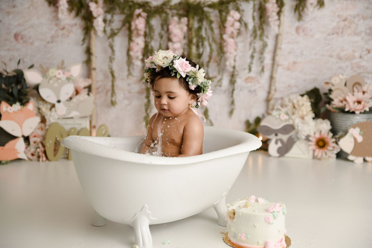 Baby girl in floral crown sitting in white bathtub during cake smash photo session.