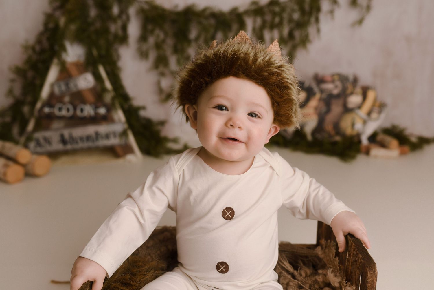 Smiling baby in cream onesie with fluffy brown hat sits in rustic wooden bowl before adventure-themed backdrop.