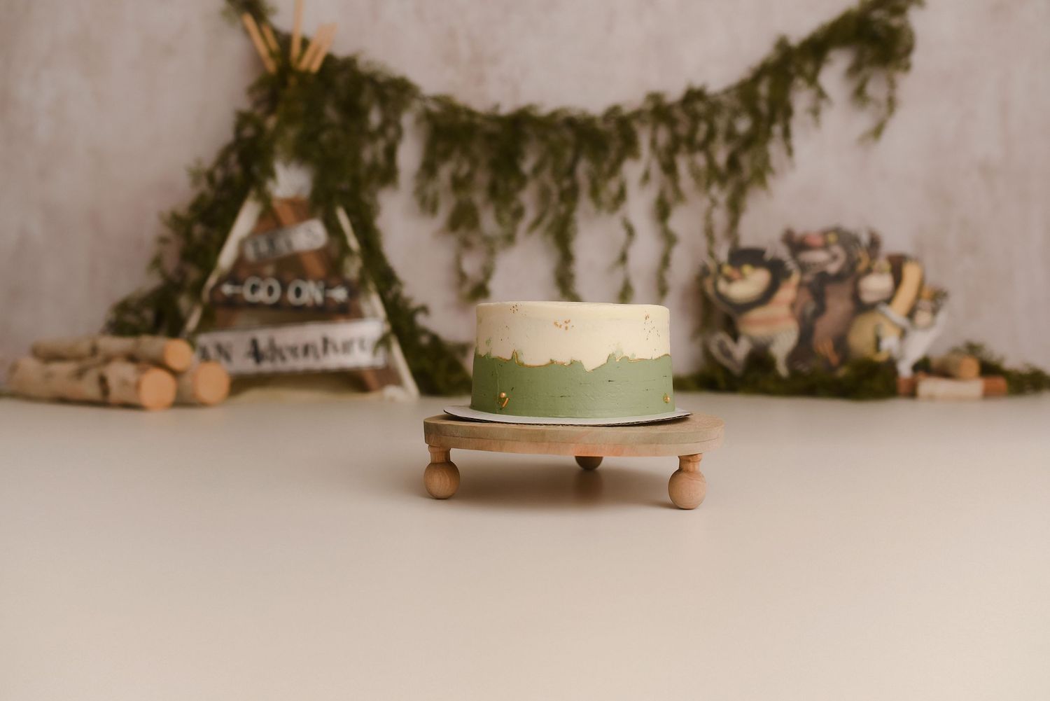 Green and white cake on wooden stand with adventure-themed backdrop featuring teepee and forest decorations.