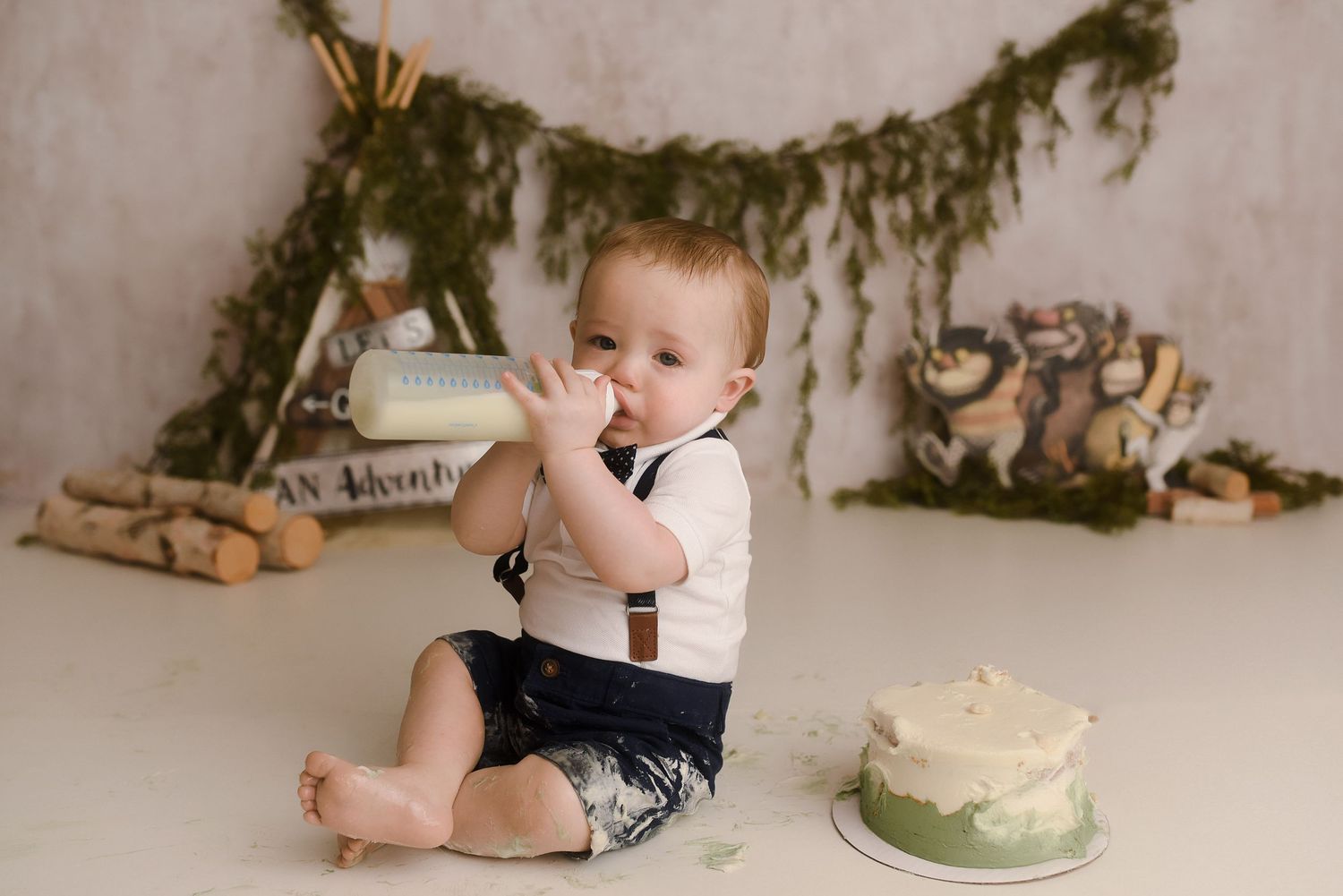 Baby boy in suspenders enjoys his first birthday cake smash session with a green woodland adventure themed setup.