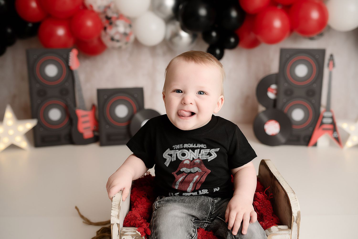 Smiling baby in Rolling Stones shirt sits in wooden box with rock music themed backdrop and red black balloons.