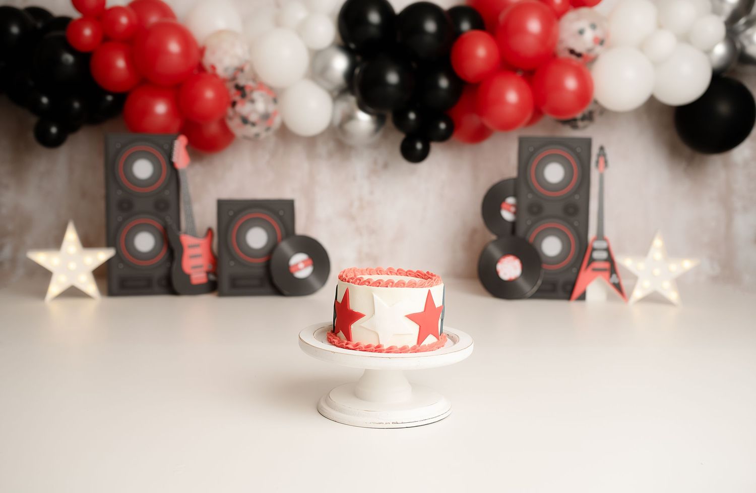 Rock and roll birthday cake smash setup with red black white balloons guitars speakers and star decorations.