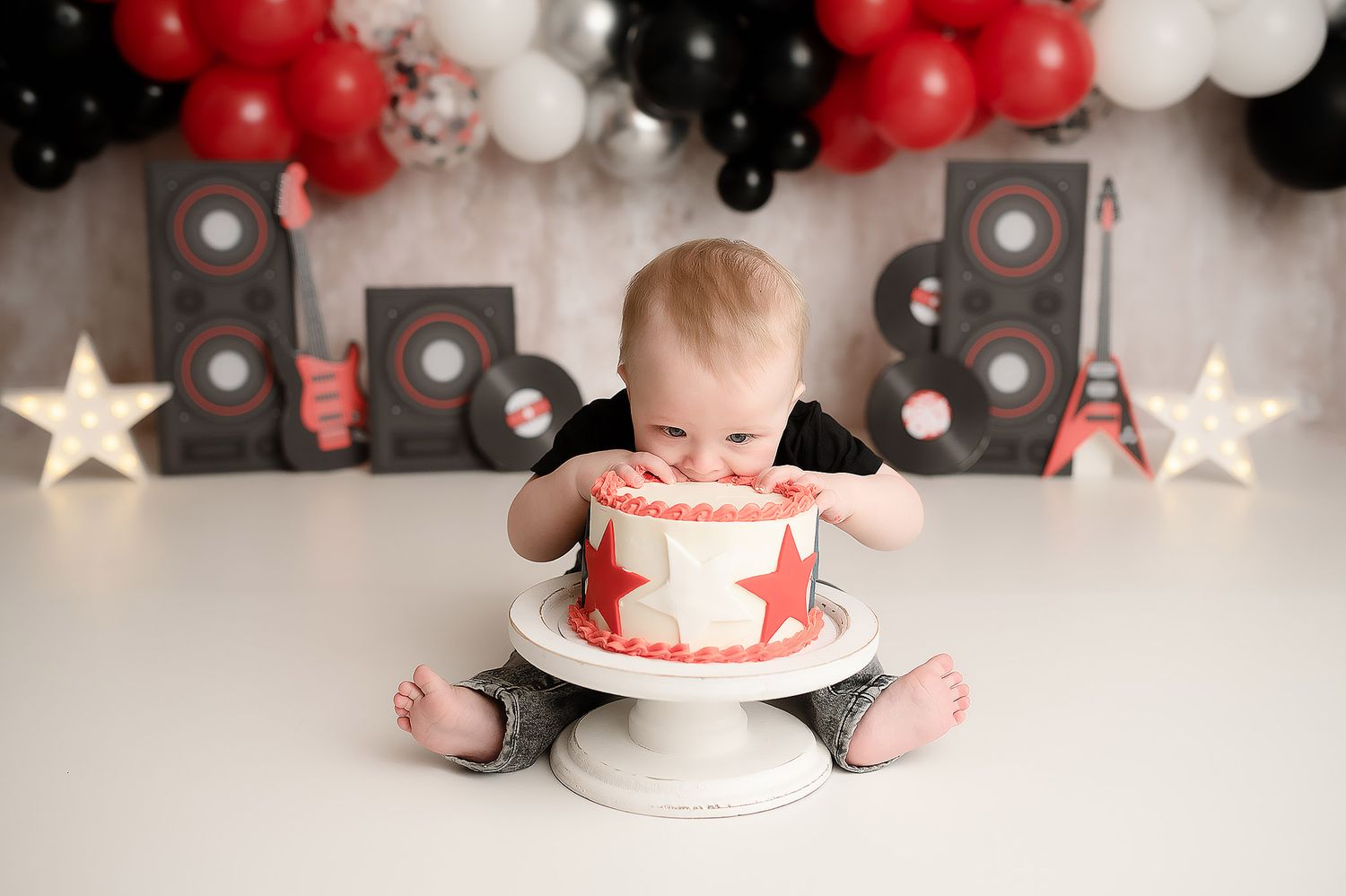 Baby smashes red and white star cake on white pedestal with rock music themed balloon backdrop.