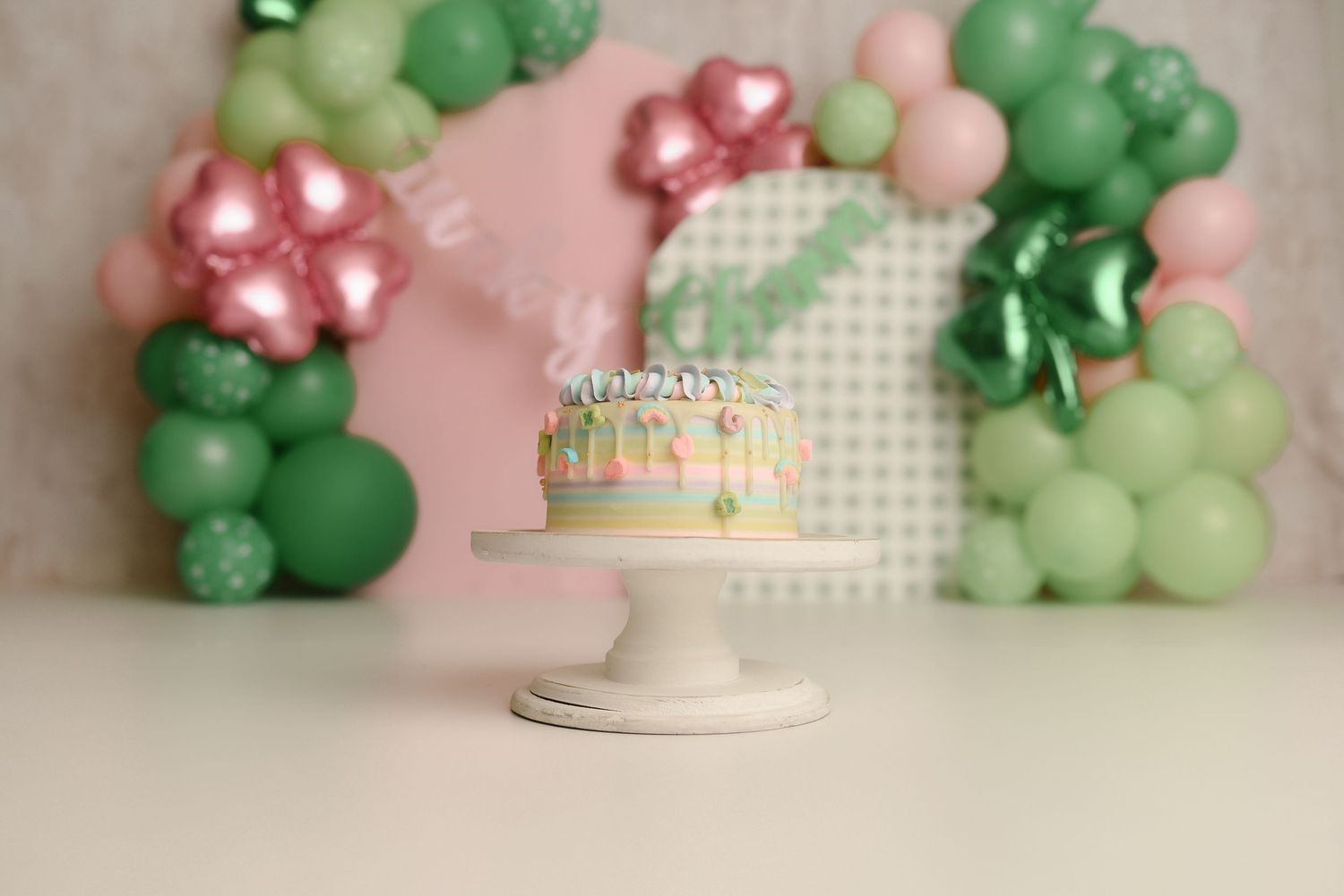 Pastel birthday cake on white stand with green and pink balloon garland backdrop for a festive celebration.
