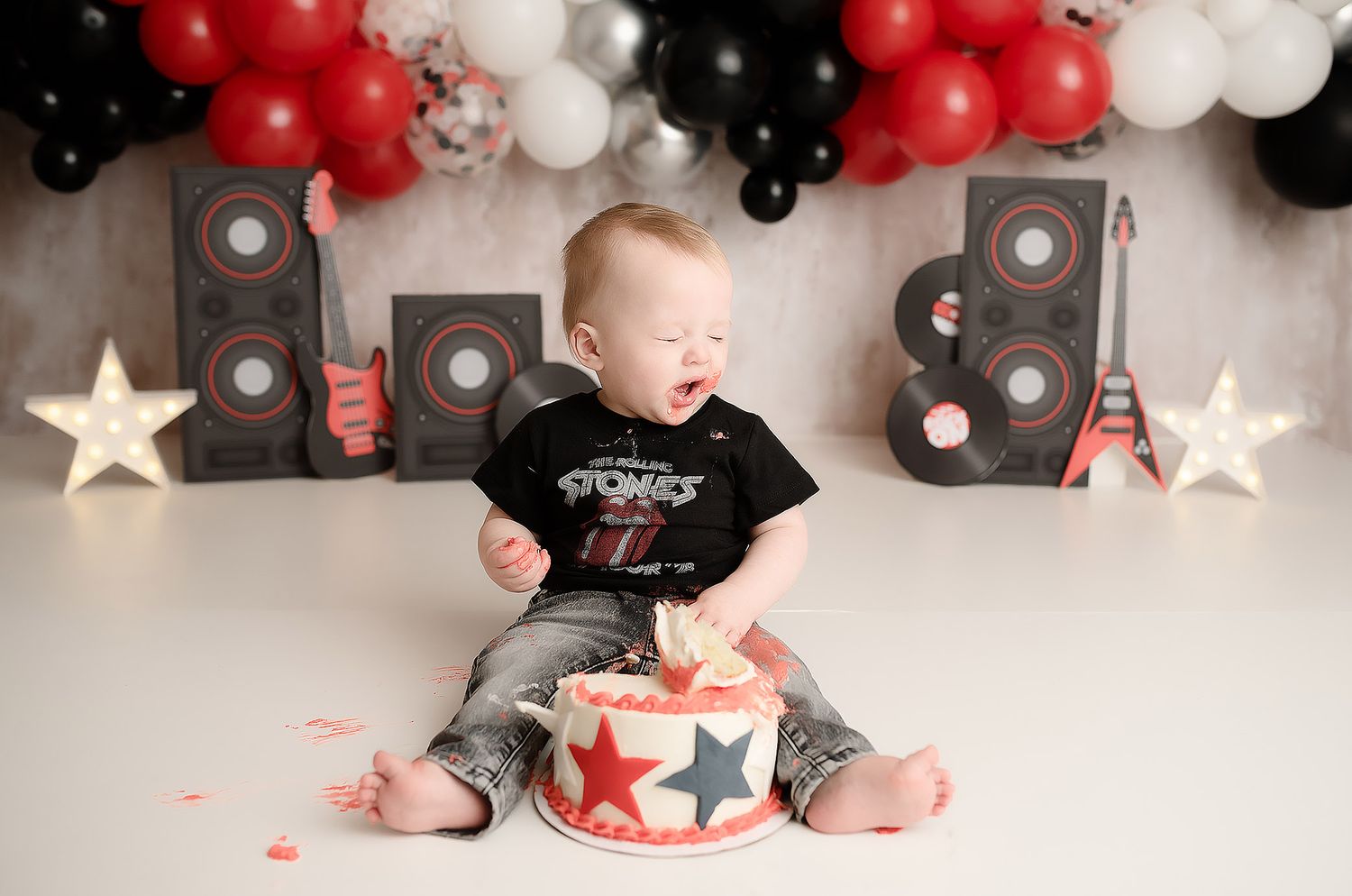 Laughing baby in Rolling Stones shirt smashes red star birthday cake with rock music balloon backdrop.