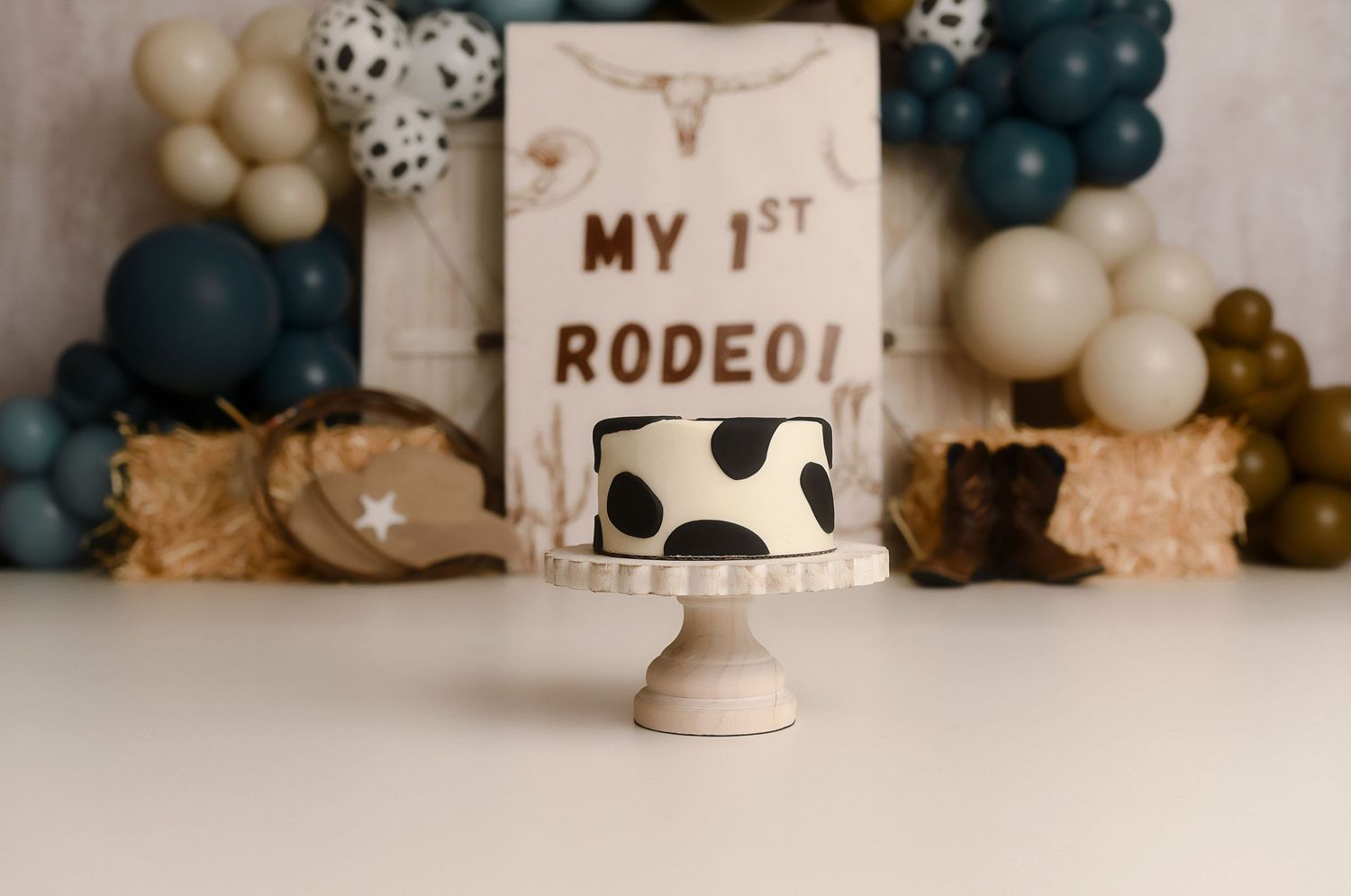 Cow print smash cake on a white pedestal stand for a baby's first rodeo birthday party with balloon backdrop.