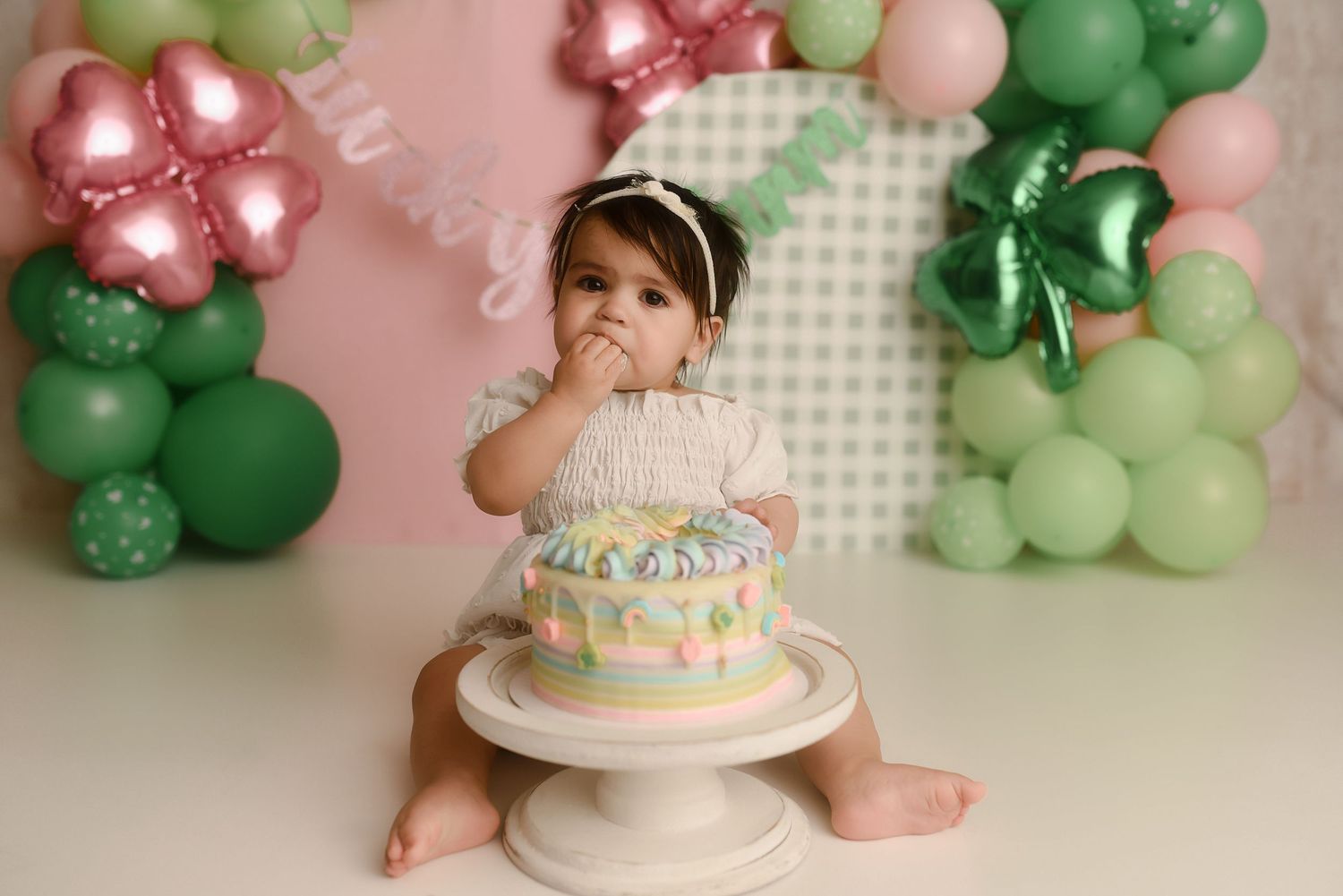 Baby girl in white dress smashes first birthday cake surrounded by pink and green balloon decorations.