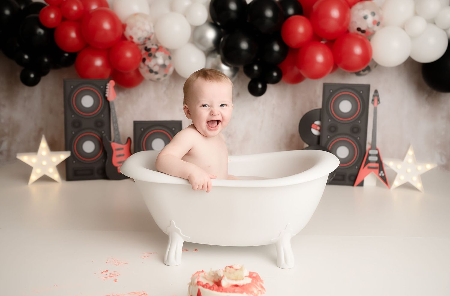 Happy baby laughs in white clawfoot tub during rock and roll themed birthday photoshoot with balloons.