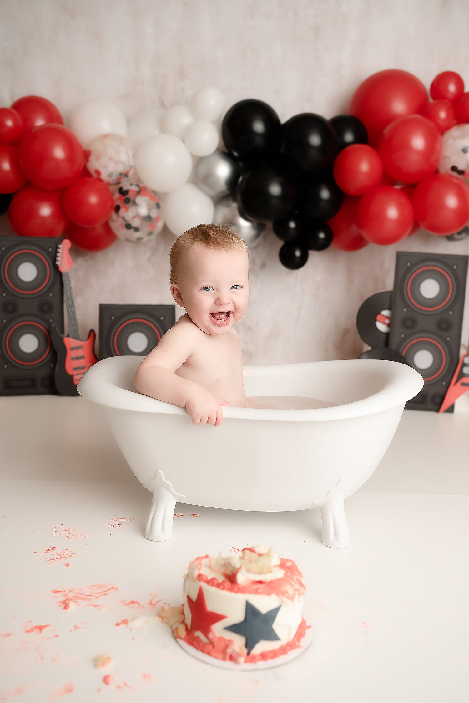 Happy baby sits in white clawfoot tub with smashed cake nearby and red black white balloon backdrop.