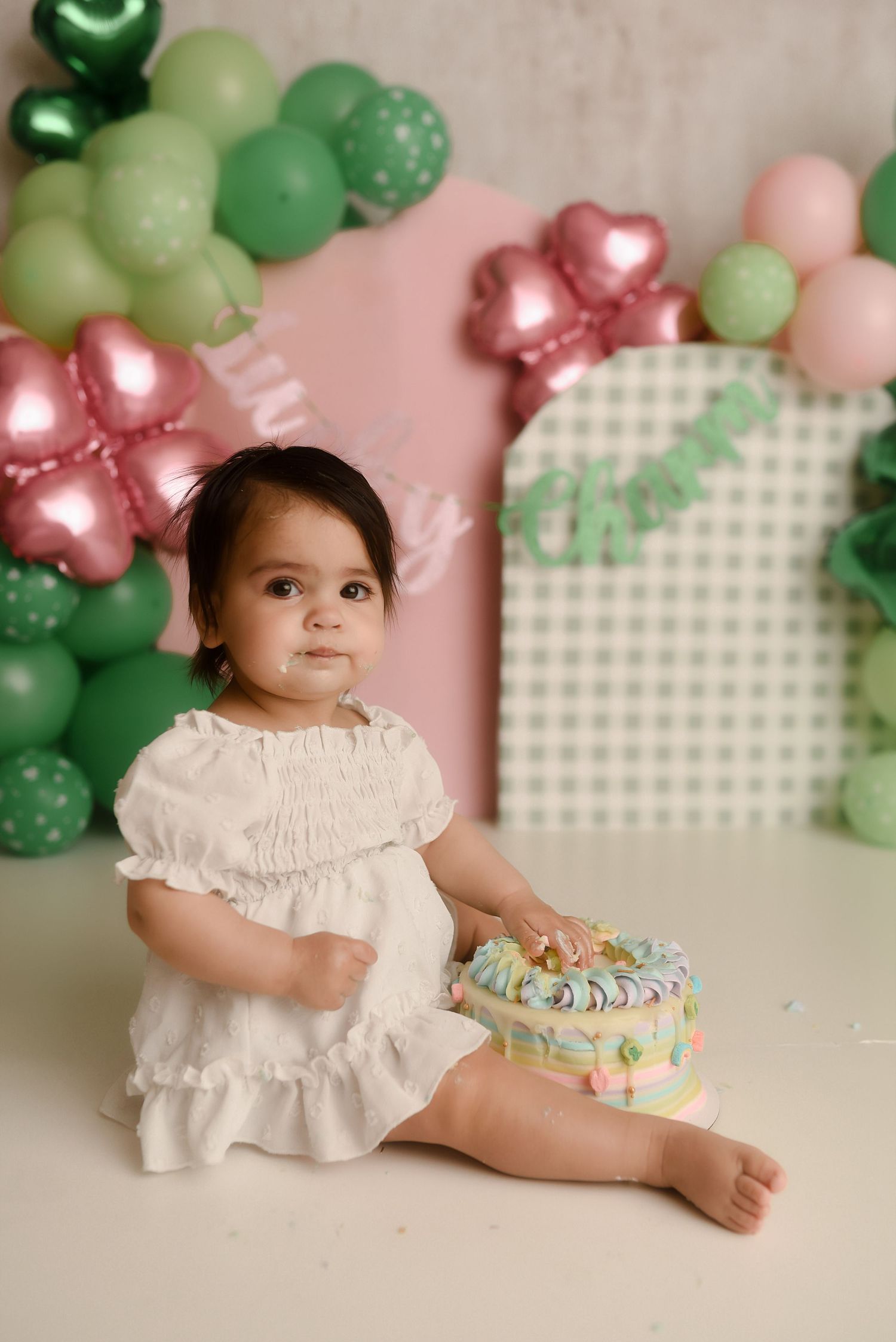 Baby girl in white dress sits with colorful birthday cake at cake smash session with pink and green balloon decorations.