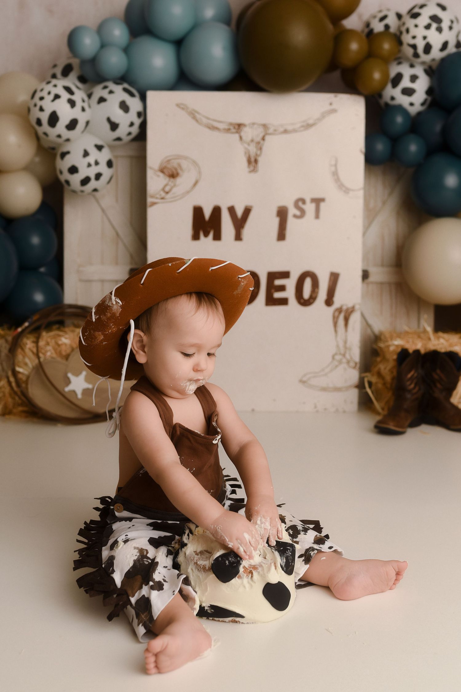 Baby in cowboy hat and cow print outfit sits before a My 1st Rodeo balloon backdrop birthday setup.