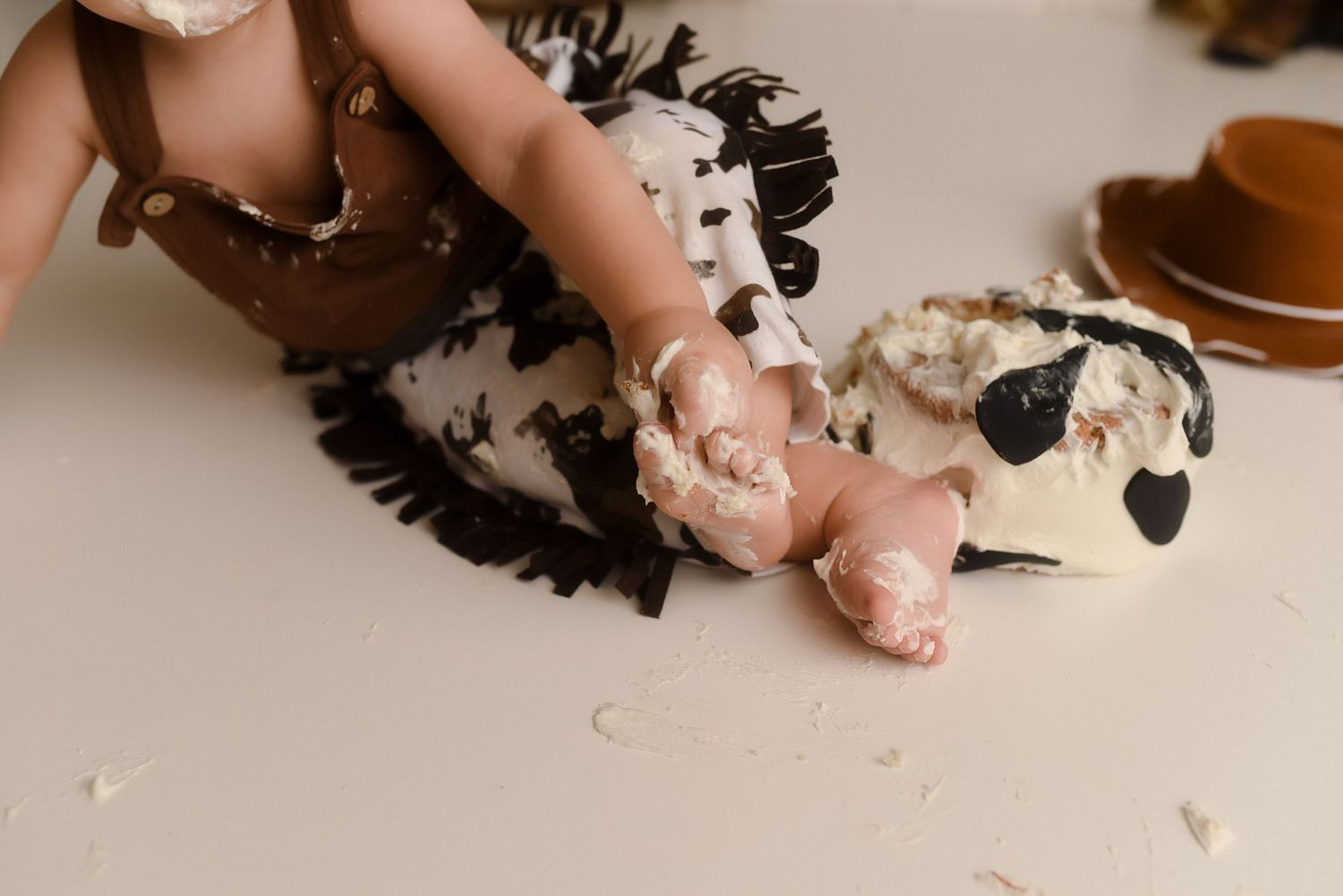 Baby smashing a cow-print cake with frosting-covered hands and feet during a first birthday cake smash photo session.