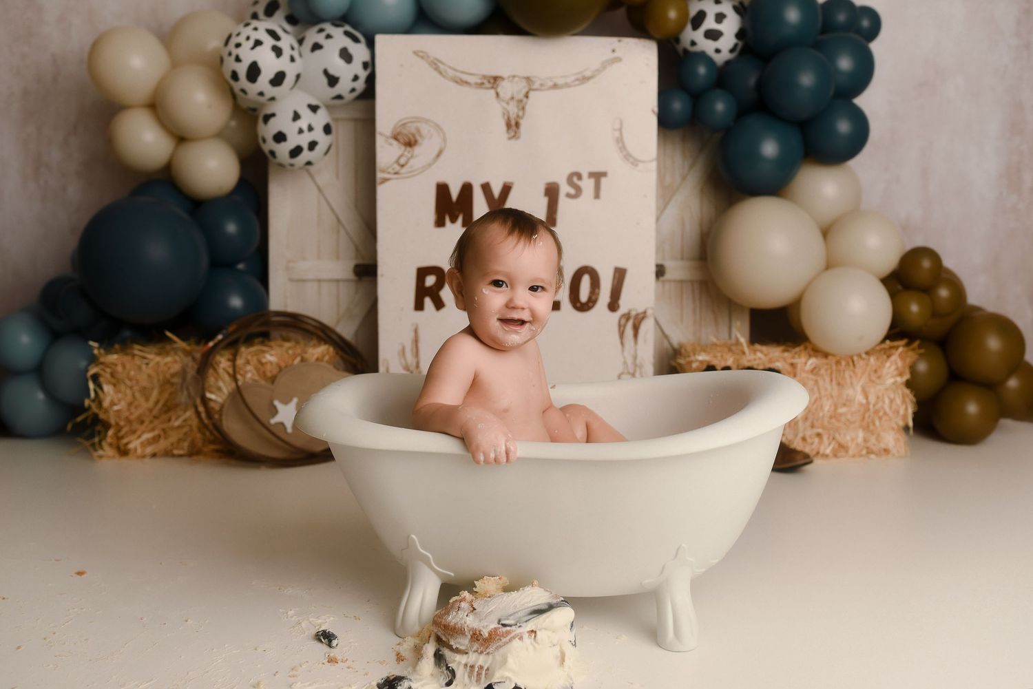 Baby smiling in white bathtub at first birthday rodeo cake smash with teal balloons and western decor.