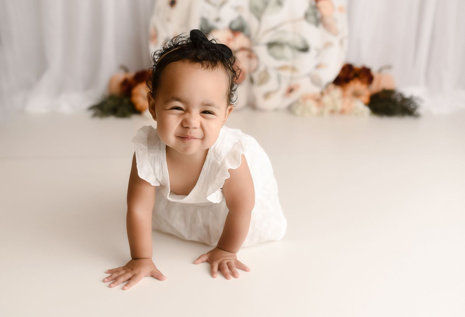 Smiling baby girl in white romper crawling on white floor with floral backdrop.