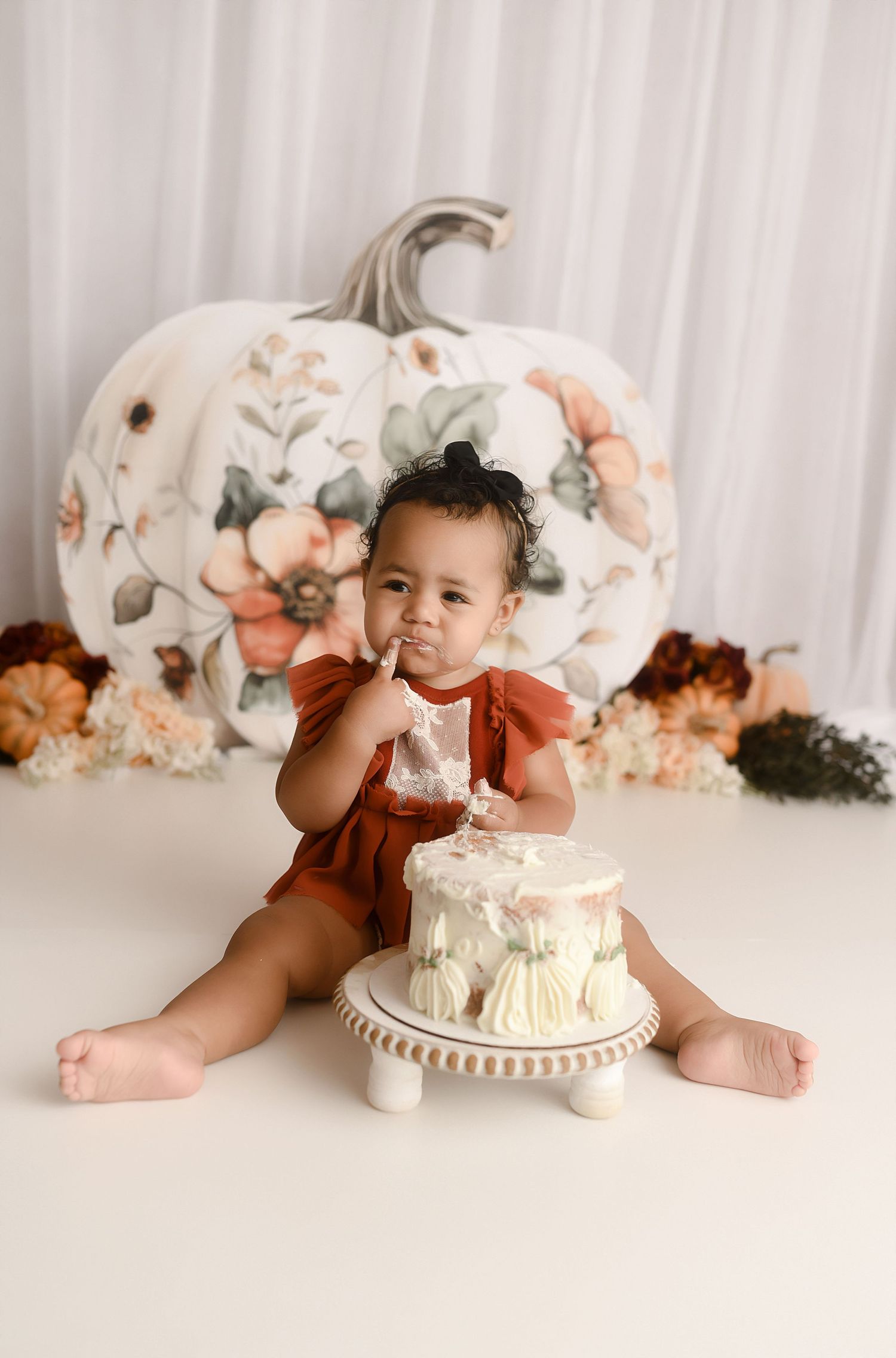 Baby girl in rust romper eating cake beside large floral pumpkin backdrop.