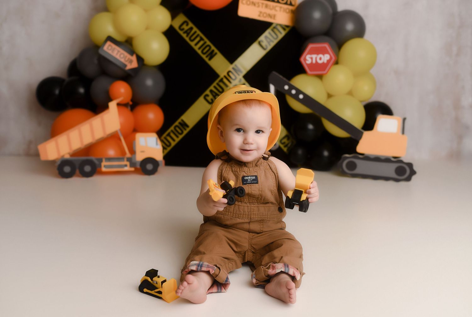 Baby in construction outfit sits before balloon backdrop with toy trucks for a themed first birthday cake smash photo.