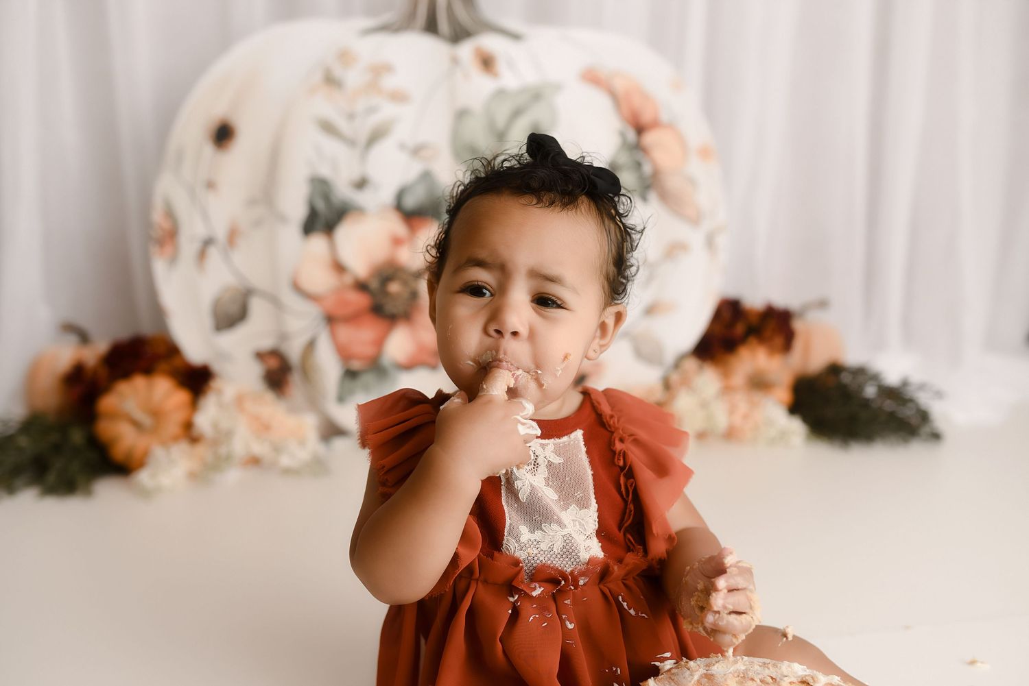 Cute baby girl in rust romper eating cake in front of floral pumpkin backdrop.