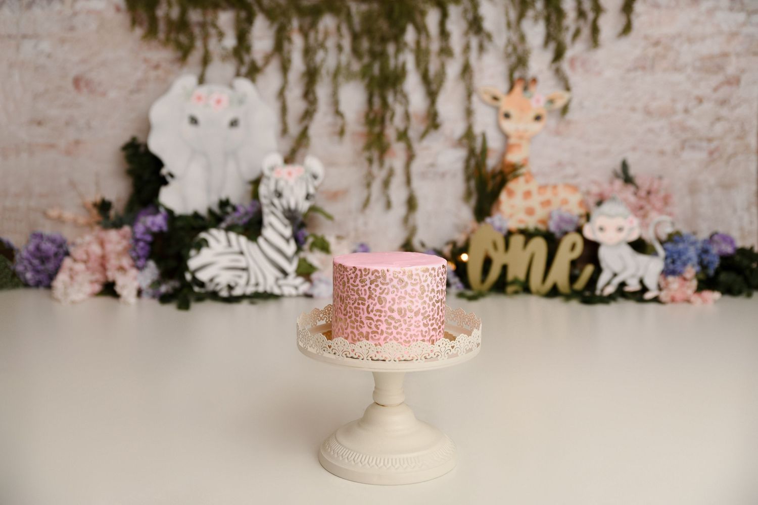 Pink floral smash cake on white stand with jungle animal decorations and one sign.