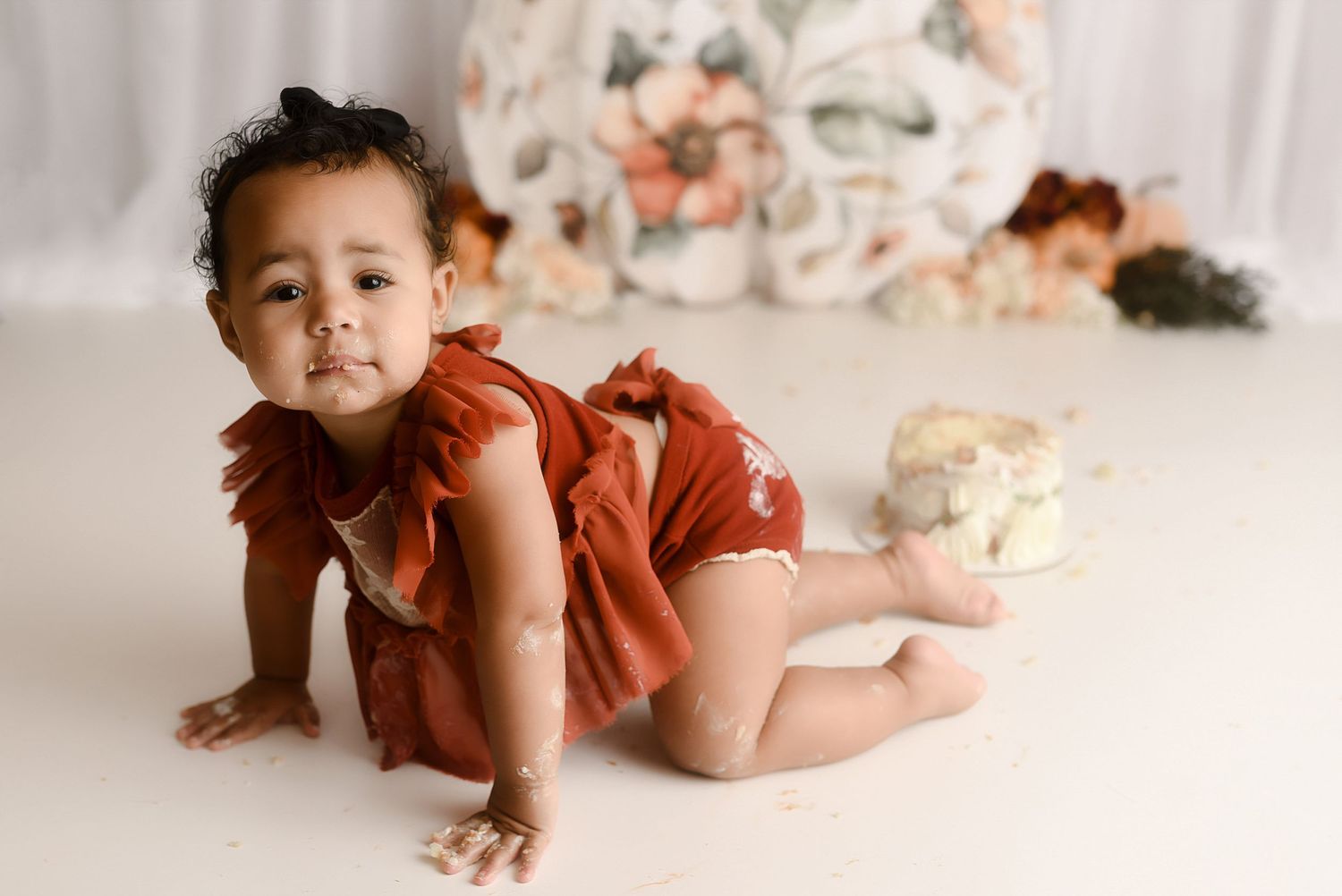 Baby girl in rust romper crawling on white floor with smashed cake in background.