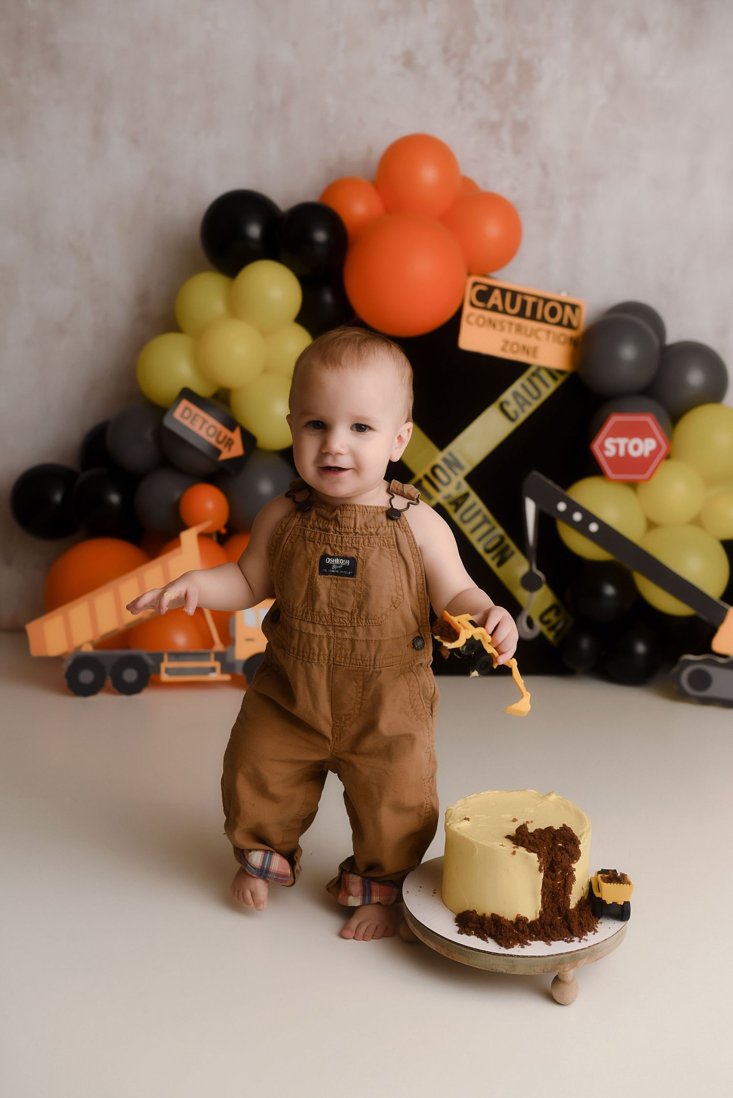 Baby boy in brown overalls smashing a construction-themed first birthday cake with balloon backdrop.