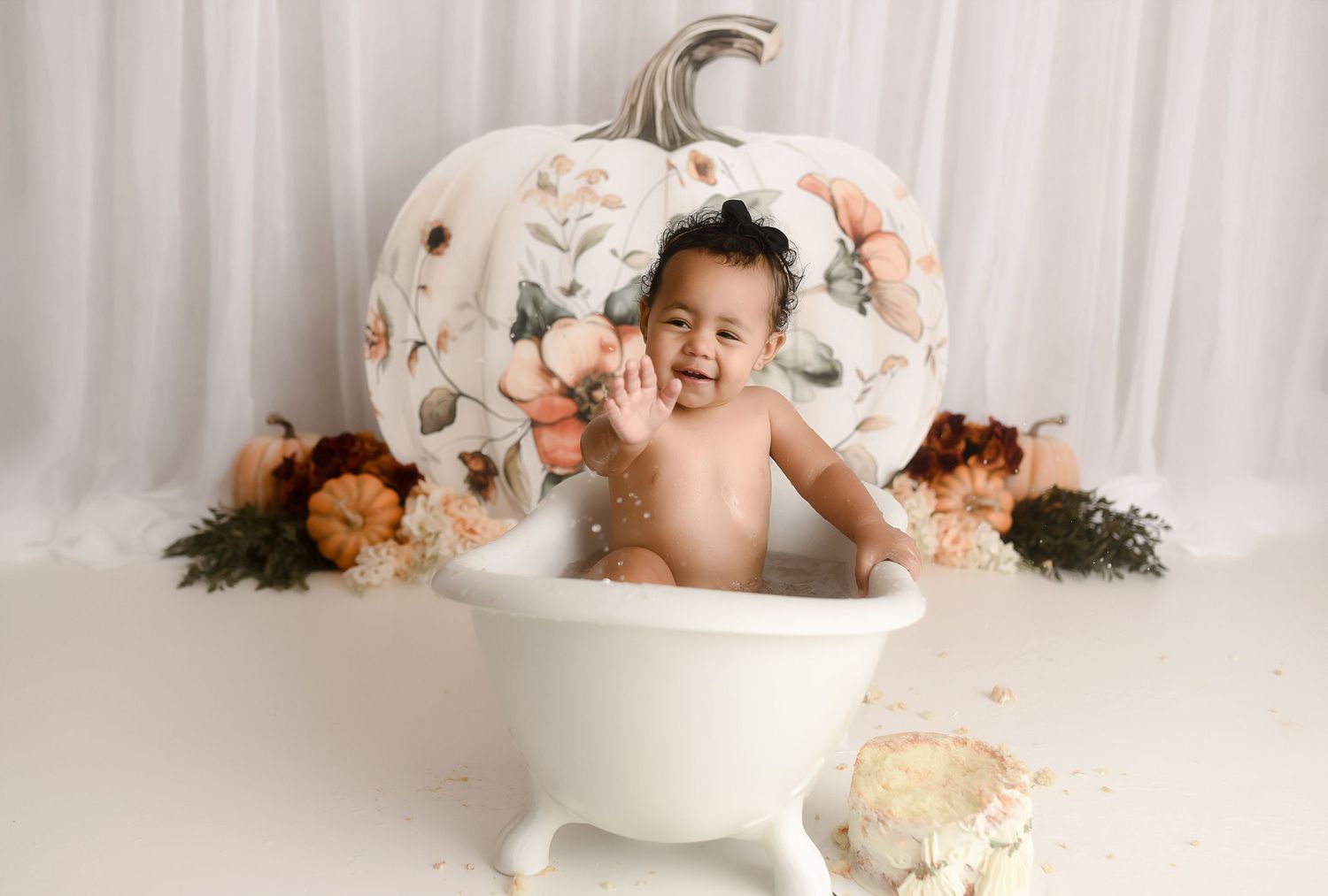 Happy baby splashing in small white bathtub with floral pumpkin backdrop.