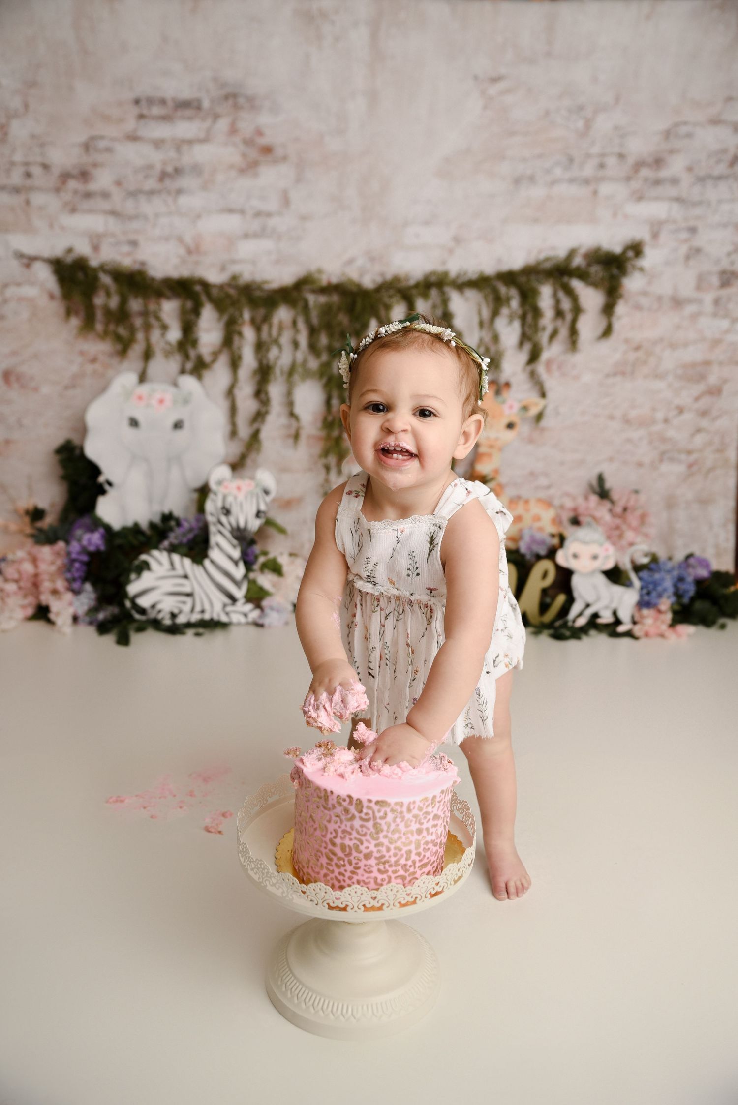 Smiling baby girl in white romper smashing pink cake at first birthday jungle theme session.